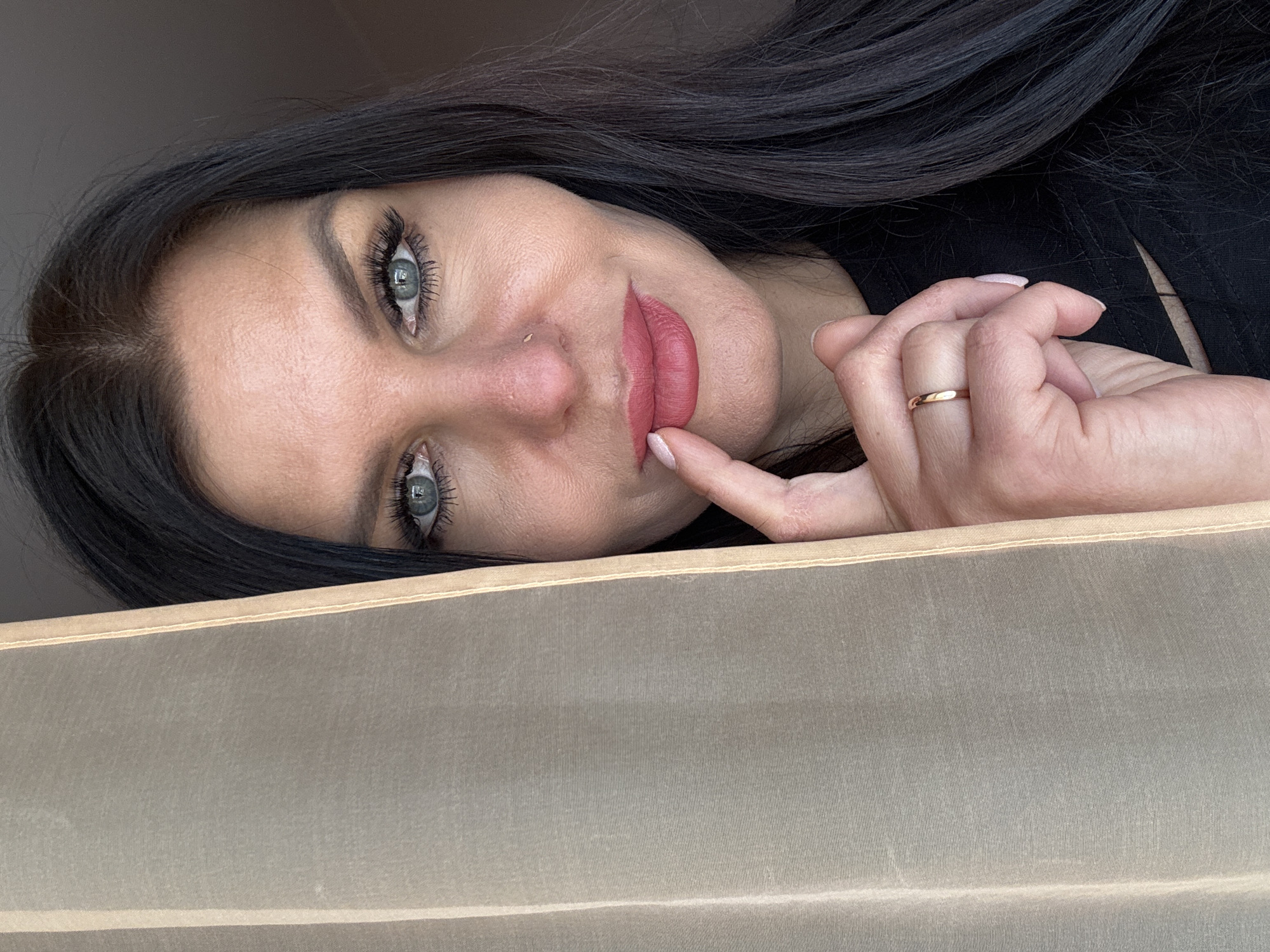 The image displays a headshot of a woman with long, dark hair and striking blue-green eyes, captured at an angle that suggests the photo is rotated counter-clockwise. She is looking directly at the viewer with a subtle, engaging smile, her lips colored with a reddish-pink lipstick. A small nose stud is visible on her left nostril.
Her right hand is raised, with her index finger gently touching her lower lip, conveying a thoughtful or playful expression. A thin, plain gold or rose gold band ring is worn on her right ring finger. She appears to be wearing a dark, possibly black, top, visible on the upper right portion of the frame.
The lower half of the image is occupied by a light beige, somewhat translucent fabric or surface, likely a curtain or blind, which adds a layered depth to the composition. The background behind the woman is a plain, light color, suggesting an indoor setting. The lighting is soft and even, emphasizing her features. There is no visible text or specific location cues beyond the general indoor environment. Her direct gaze and hand gesture create an intimate and captivating mood.