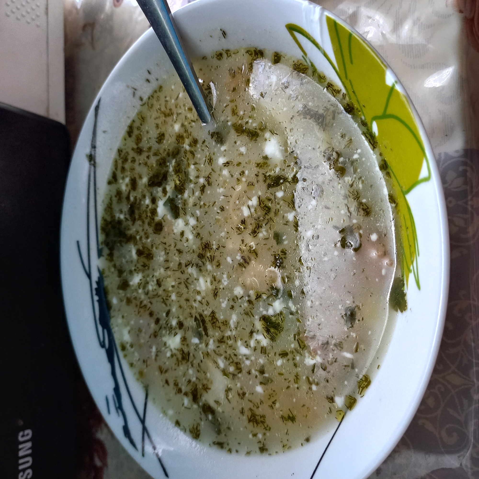 A close-up shot focuses on a bowl of soup. The soup appears to be broth-based with visible pieces of meat and finely chopped green herbs, possibly dill, sprinkled throughout. Small white flecks, likely from cheese or solidified fat, are also present. A silver spoon is partially submerged in the soup on the right side of the frame. The bowl itself is white with a decorative pattern of blue lines and green leaf shapes along the rim. The background is out of focus, showing a dark object with the word "SUNG" visible on it, suggesting it might be an electronic device. The surface the bowl rests on has a patterned tablecloth. The scene is indoor, likely a kitchen or dining area. There are no people visible in the image. The location is identified as Klyntsi, Ukraine.