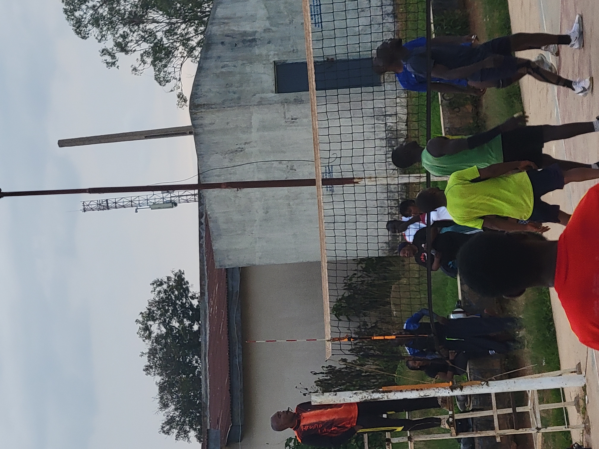 The image depicts a group of individuals engaged in a volleyball match outdoors. In the foreground, a man in a red shirt is partially visible from behind, facing away from the camera. To the left, a man in an orange and black shirt stands on a ladder, looking towards the left side of the frame. A volleyball net stretches across the midground, with several players on both sides. To the left of the net, a woman in a black shirt and another person in a blue shirt are visible. To the right of the net, several men are playing, including one in a green sleeveless shirt, one in a blue shirt, and another in a dark blue shirt and shorts. Behind the players, a chain-link fence encloses the court. In the background, there is a building with a metal roof and a concrete wall, along with trees and a telecommunications tower. The sky is overcast, suggesting a cloudy day. The scene appears to be an informal sporting event taking place in an urban or semi-urban environment.