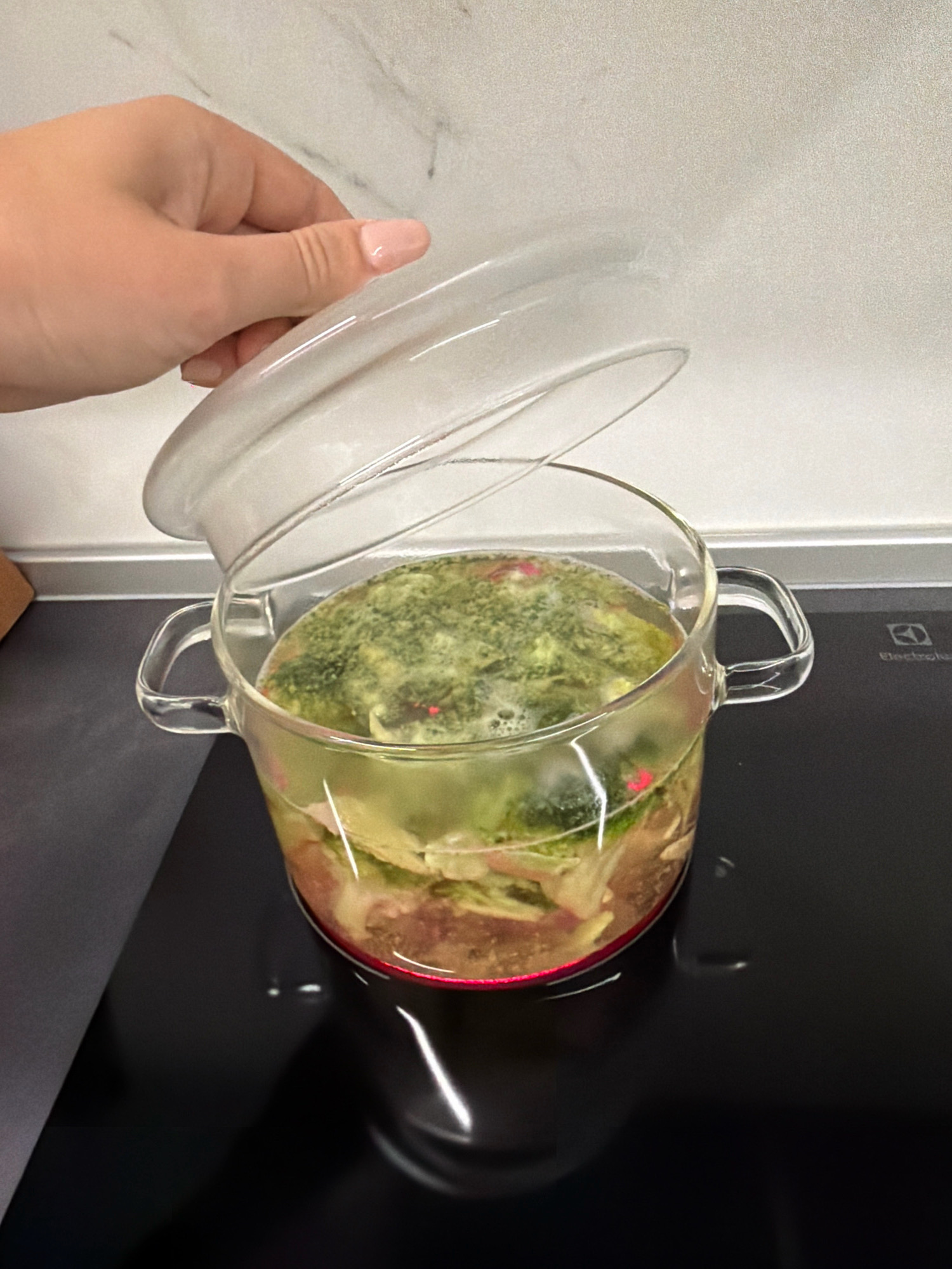 A person's hand is lifting the glass lid off a clear glass pot filled with a green soup, which is placed on a black induction stovetop. The soup appears to contain pieces of vegetables and possibly meat. The background shows a white marble-patterned backsplash. The scene is indoors, likely in a kitchen. There are no specific indicators to place the location in Sofia, Bulgaria.