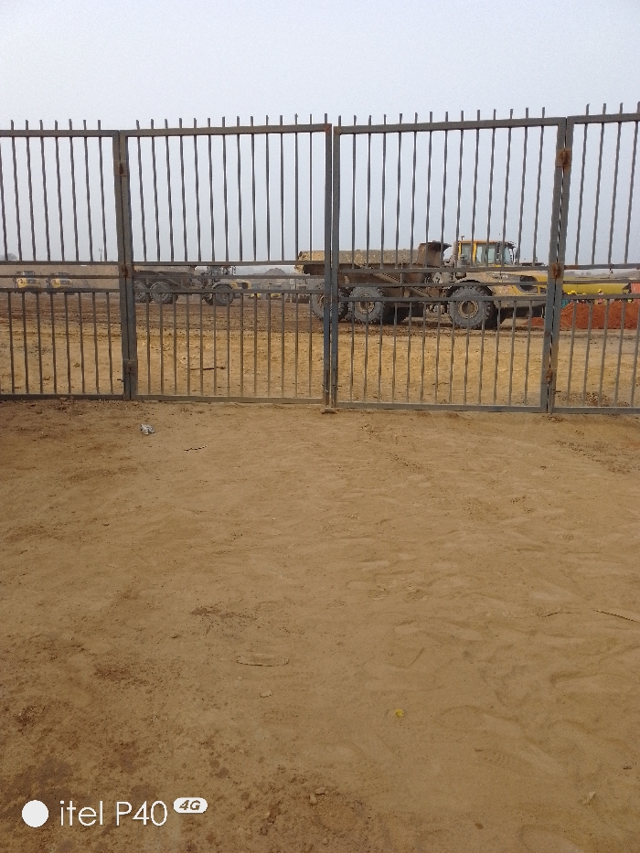 A fenced area of sandy ground is seen from behind a gate, revealing a construction site. Large yellow machinery, possibly dump trucks, are visible through the bars of the gate. The sky is overcast, suggesting a dull day. The ground in front of the gate is bare and sandy, likely the same material found within the fenced area. A logo from a phone is partially visible in the corner.