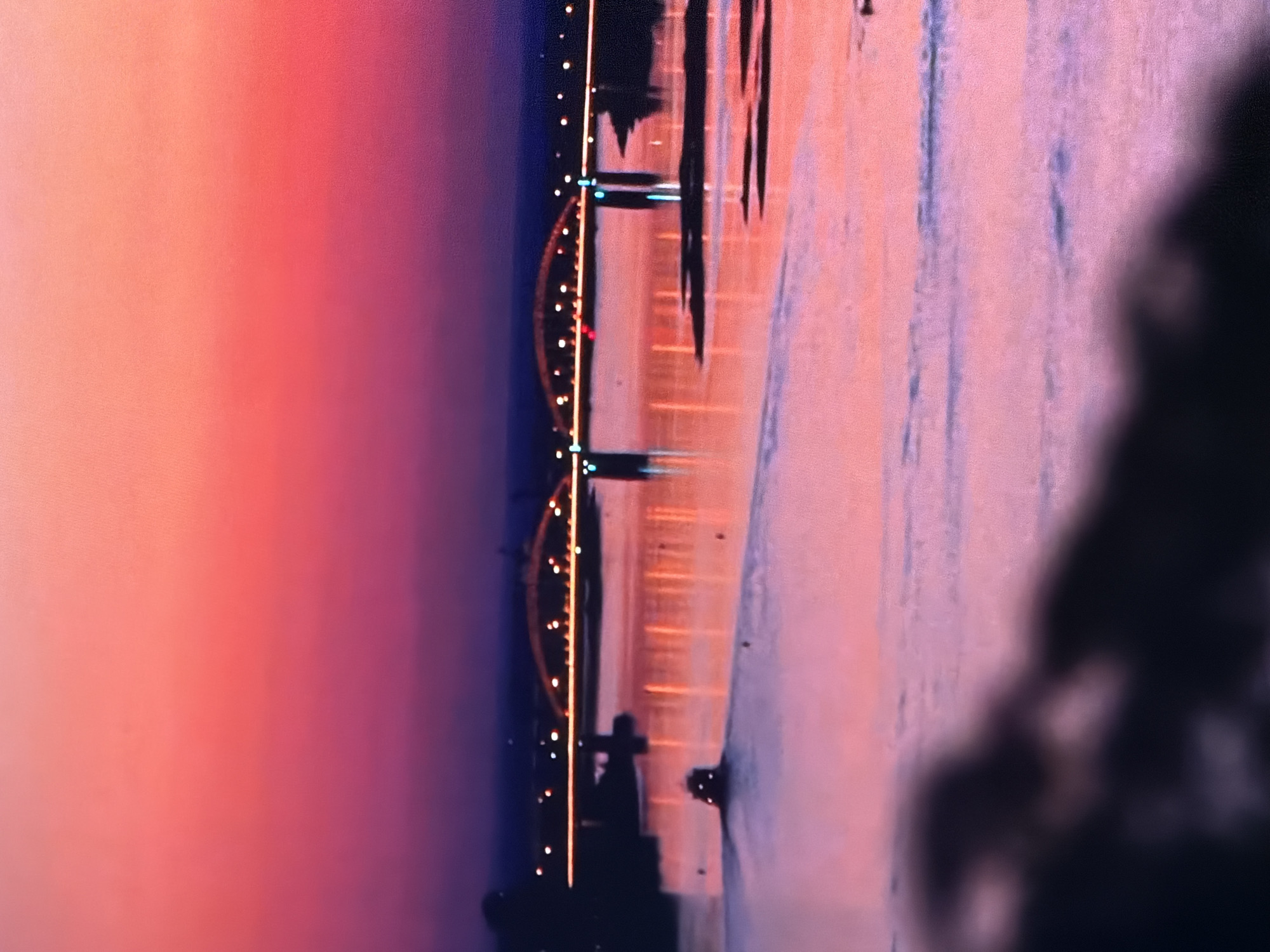 An expansive, blurry landscape captures a body of water reflecting the vibrant colors of a sunset. The sky transitions from a bright pink at the top to a deep purple near the horizon. A bridge with illuminated arches spans the water in the mid-ground. On the water, a small boat with its lights on is moving away from the viewer, leaving a wake. The foreground is dominated by a dark, out-of-focus mass, likely land or foliage. The overall scene is characterized by its soft focus and the striking color palette of the sunset.