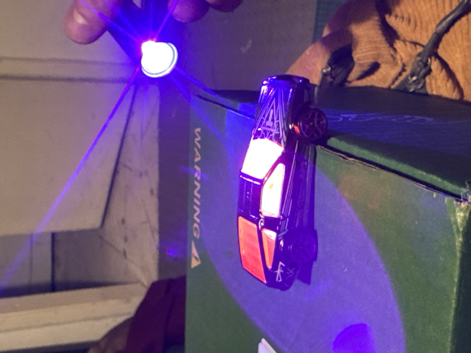 A brightly lit toy car with white and red lights sits atop a dark green box labeled "WARNING." A hand holds a small, white light source above the car, casting a purple glow on the car, the box, and the surrounding environment. The background includes part of a white shelf, a brown garment, and a black wire. The location is not discernible from the image.