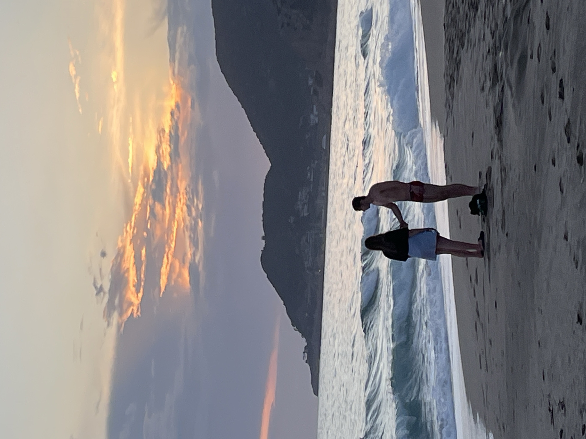 The image shows two people standing on a beach in Alanya, Turkey, at sunset. One person, male, is wearing red swim trunks. The other person, appears to be female, is wearing a black top and light-colored shorts. They are facing the ocean. The sea is moderately rough with visible waves. In the background, there's a large mountain. The sky is filled with clouds colored orange and yellow from the setting sun. The beach sand is light-colored and appears wet.
