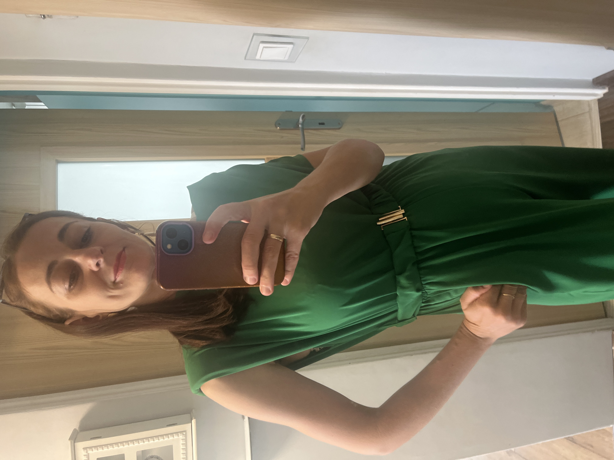 A woman stands in a room, taking a selfie in a mirror. She is wearing a green sleeveless dress with shoulder pads, a gold belt, and a gold ring on her left hand. Her right hand is in her pocket. She has brown hair and is wearing glasses on top of her head. The room appears to be a bathroom or dressing area, with a door and a light switch visible in the background.