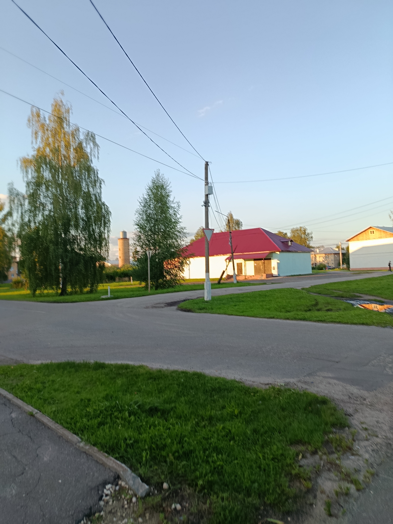 The image depicts a street scene in Veriya, Russia, at what appears to be late afternoon or early evening. The sky is clear and light blue, with a few wispy clouds. Power lines stretch across the upper portion of the frame. In the middle ground, there is a road intersection with green grass verges. On the right side of the road, a white building with a red roof is visible, and further to the right, another building with a cream-colored facade and a darker roof. A lone figure, partially obscured, is walking on the sidewalk in front of the cream-colored building. To the left of the road, a tall, cylindrical, brick structure, possibly an old water tower, is visible in the distance behind a cluster of trees. Several trees with green foliage are present throughout the scene, with reflections of their leaves visible on the power lines. The ground is paved with asphalt on the roads and sidewalks, and the grass is well-maintained. The overall impression is one of a quiet, residential area.