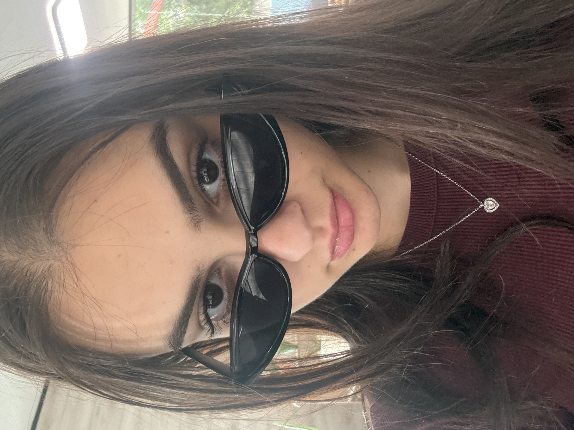A young woman with dark hair and fair skin is wearing sunglasses. She is also wearing a maroon shirt and a silver necklace with a heart-shaped pendant. The location in the city of Blagoevgrad, Bulgaria, cannot be determined from the image.