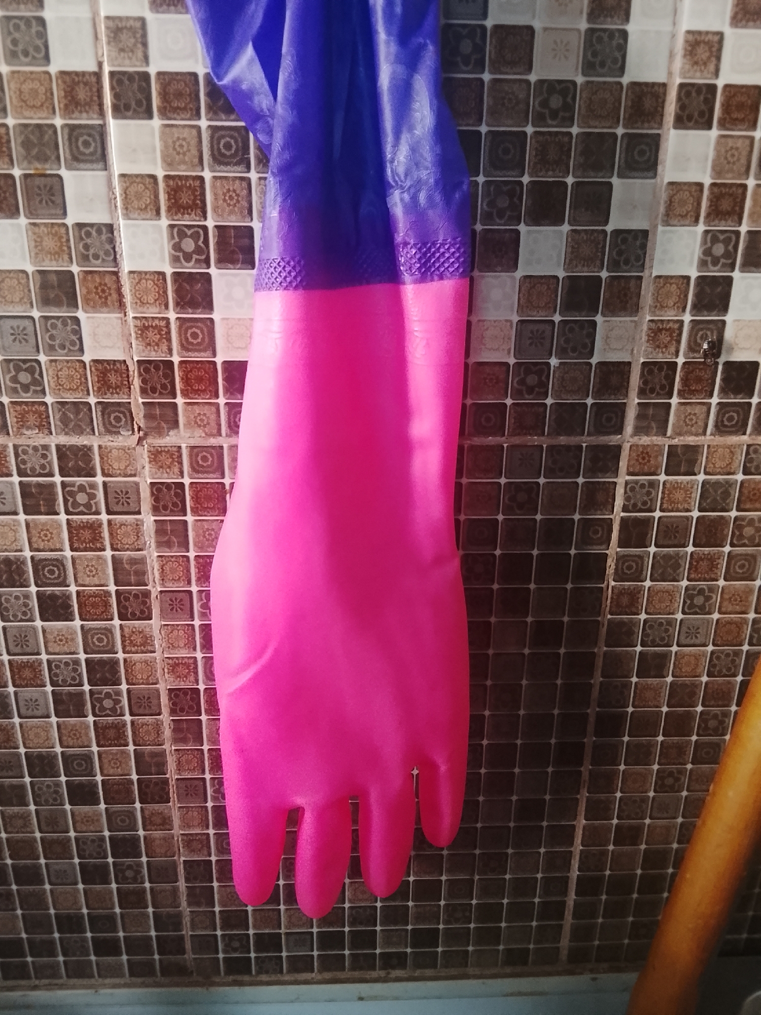 A close-up shot shows a pink and purple rubber glove hanging against a tiled wall. The glove is hanging by its cuff, with the pink portion occupying the majority of the frame, displaying the shape of fingers. The purple portion of the glove is visible at the top. The wall is covered in small square tiles with a mosaic pattern, featuring various floral and geometric designs in shades of brown, white, and gray. To the right of the glove, a portion of a wooden object, possibly a rolling pin or broom handle, is visible. The scene appears to be indoors, likely in a kitchen or utility area.