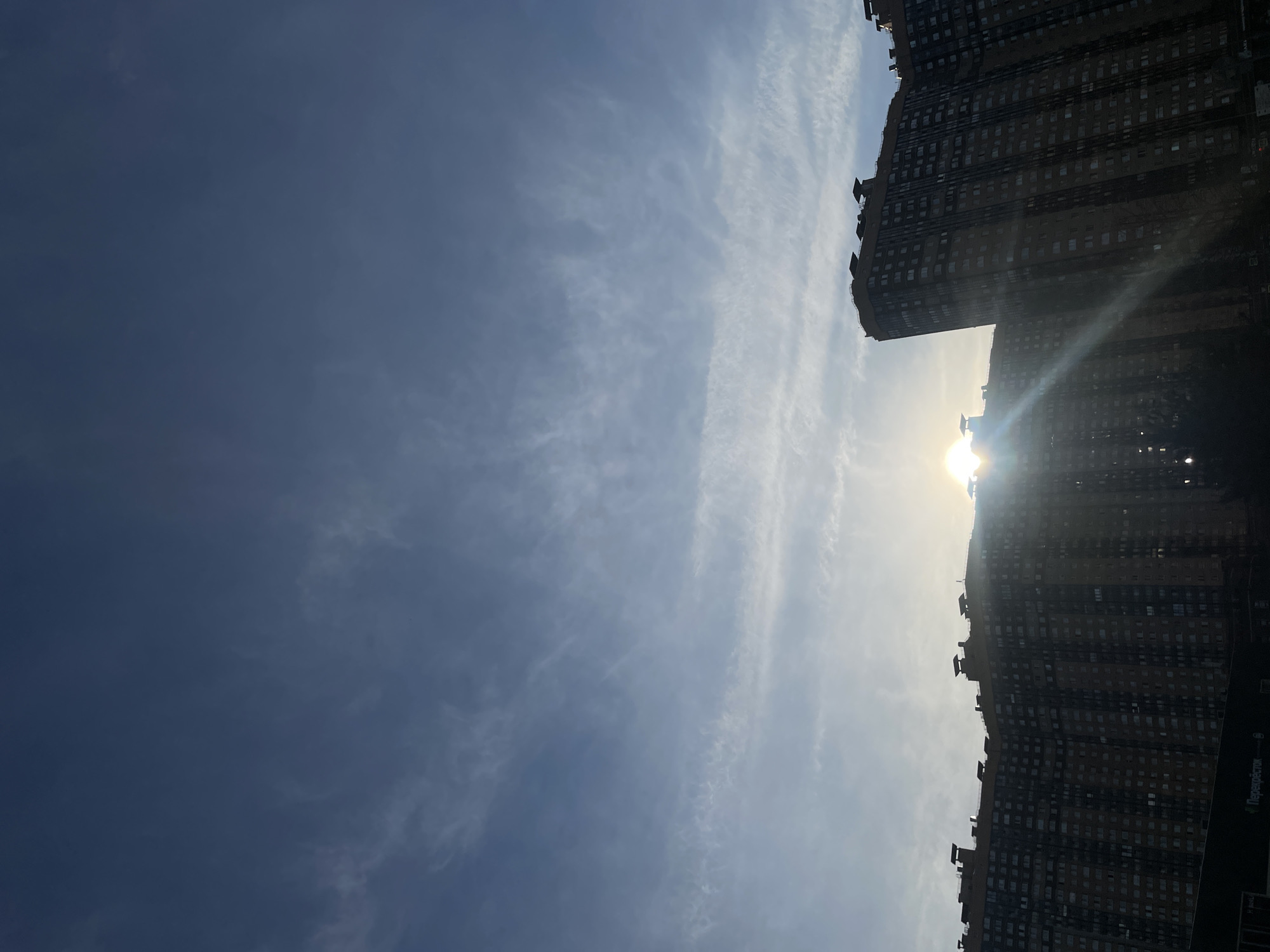 The image shows a silhouette of tall apartment buildings against a hazy sky with the sun setting. The buildings are dark and appear to be modern, with many windows. The sky is a deep blue with streaks of clouds. The sun is a bright, glowing orb peeking through the clouds and casting rays of light. The lower part of the image shows the dark silhouettes of more buildings and possibly trees, with a faint glimpse of a store sign that reads "Перекресток," a common Russian supermarket chain. The overall impression is one of an urban landscape at dusk.