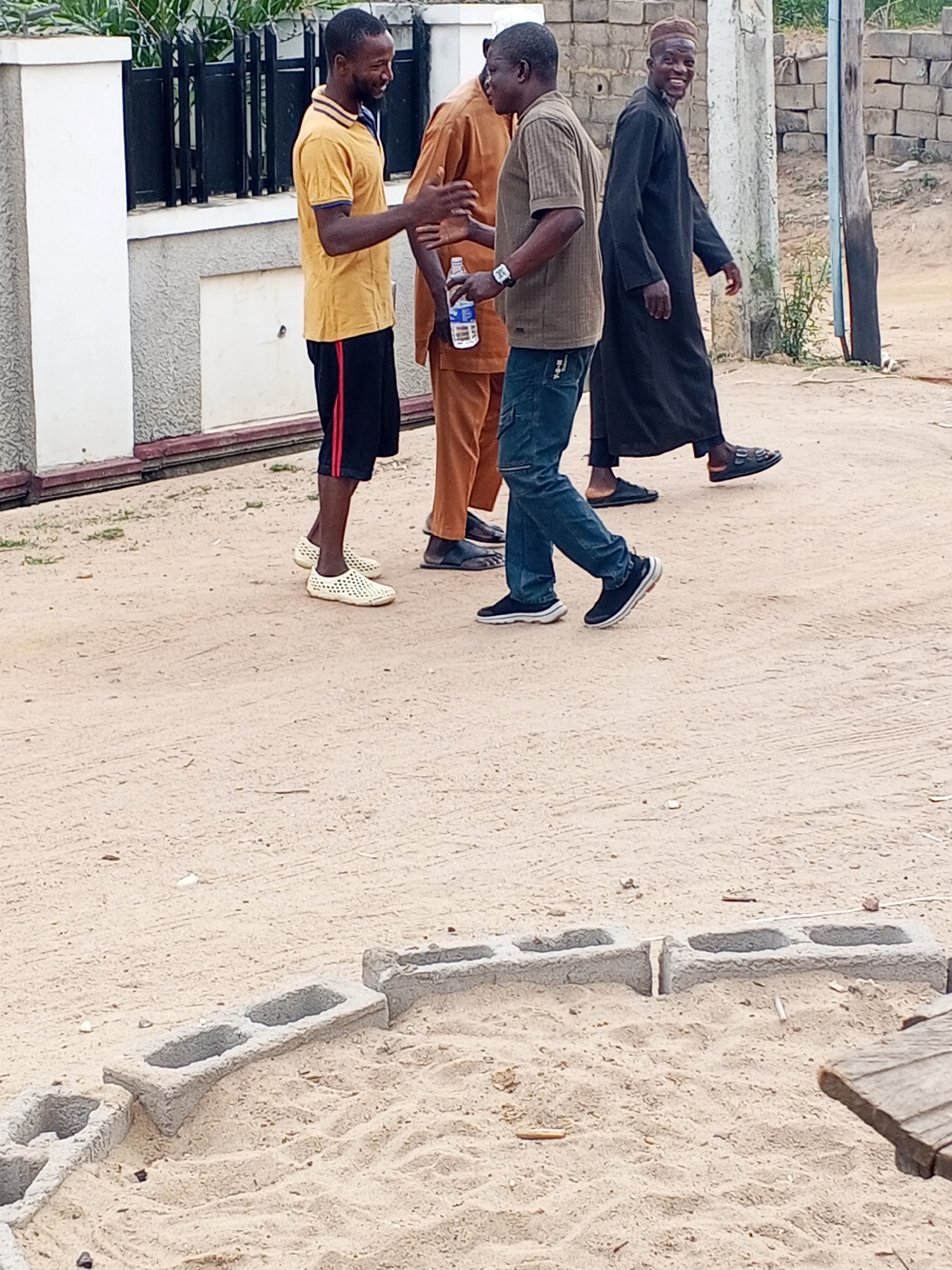 The image depicts four men in an outdoor setting in Adonkolo, Nigeria. The environment is characterized by a sandy, unpaved ground with concrete blocks arranged in a semi-circular pattern.
The first man, on the left, wears a yellow polo shirt with blue trim, black shorts with red stripes, and perforated white shoes. He appears to be in the midst of a greeting gesture with another man. The second man, wearing a brown traditional outfit, has a water bottle in his hand. The third man is dressed in a khaki short-sleeved shirt and blue jeans, walking while holding a bottle. The fourth man, on the right, is wearing a long black robe and a cap, smiling and looking toward the camera. He wears black sandals.