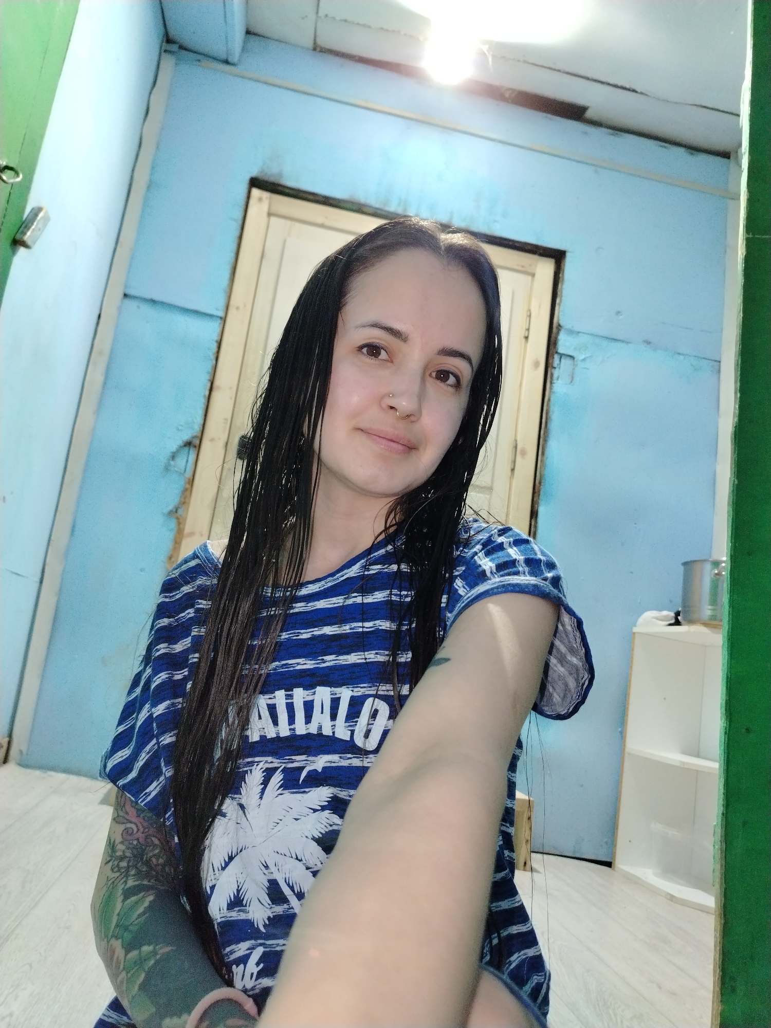 The scene is an interior room within a city of Unknown, Unknown. A light-skinned female individual, approximately 20-35 years of age, with long, dark, wet-appearing hair, is depicted from the mid-torso upward, looking directly into the camera with a slight smile. She exhibits a small ring piercing on her right nostril and wears a blue and white horizontally striped t-shirt adorned with a white palm tree graphic and the word "ATTALO". The subject's left arm displays a multi-colored tattoo sleeve, primarily green, while a small dark tattoo is visible on her upper right arm. A pink wristband is present on her left wrist. The subject's right arm is extended into the foreground, partially obstructing the view. The background features light blue paneled walls, exhibiting wear, paint deterioration, and exposed seams. A closed light wood-colored door with a dark frame is visible behind the subject. An exposed bright light source is mounted on the ceiling. To the right of the subject, a white, open-shelf unit is present, with a small, metallic silver pot resting on its top surface. The floor is covered with light-toned laminate flooring. Green trim elements are observed vertically on the extreme left and right borders of the frame.