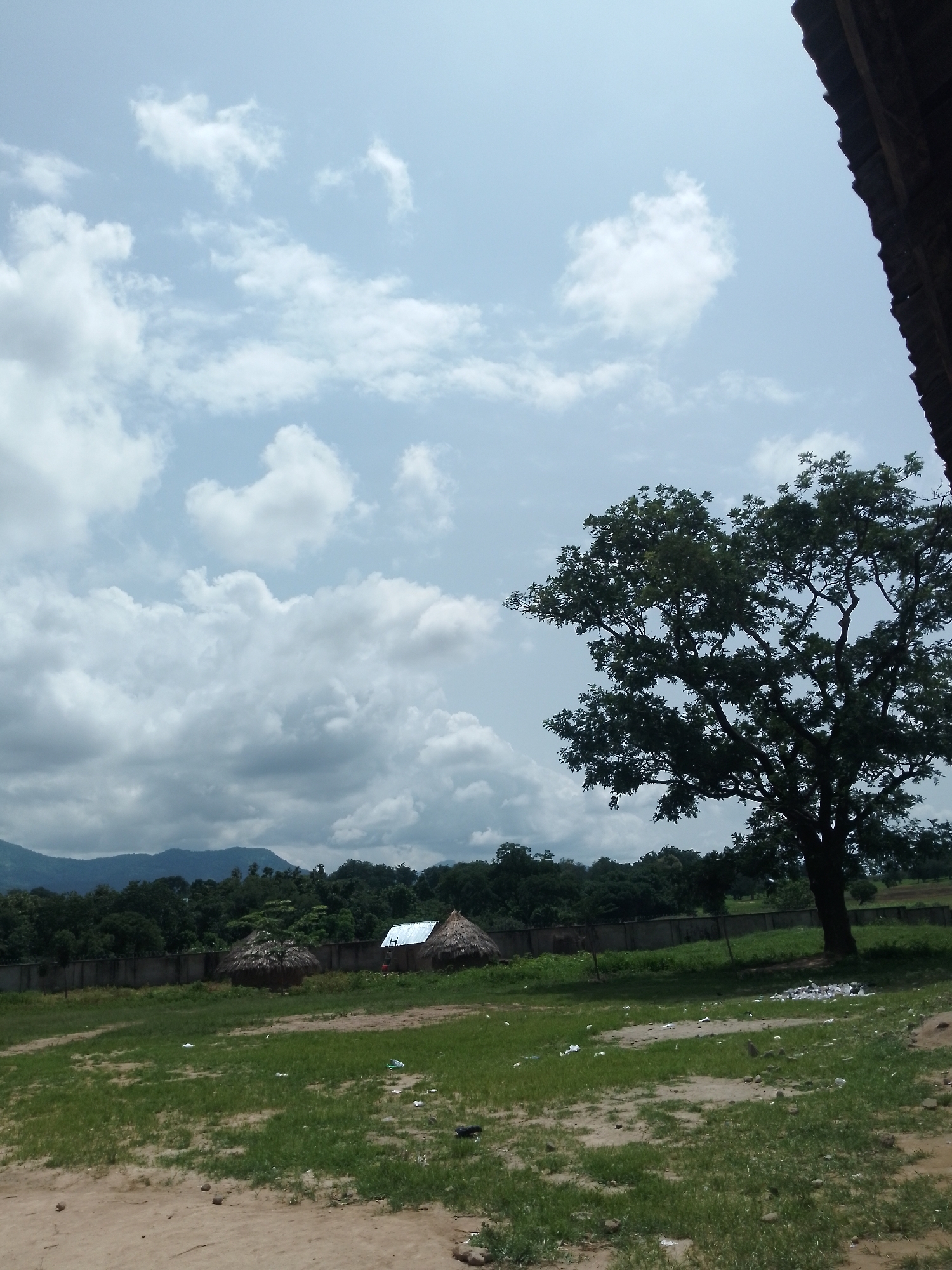 The image depicts an outdoor scene, likely in the city of Serti, Nigeria. In the foreground, a dirt path transitions into a grassy field with scattered debris. Further back, a wall with a fence separates the field from a collection of structures. Among these structures are two thatched-roof huts and a building with a metal roof. A large, mature tree with dense foliage stands prominently in the mid-ground, silhouetted against a sky filled with white and grey clouds. In the background, rolling hills covered in lush green vegetation rise against the horizon. The overall environment suggests a rural or semi-rural setting.