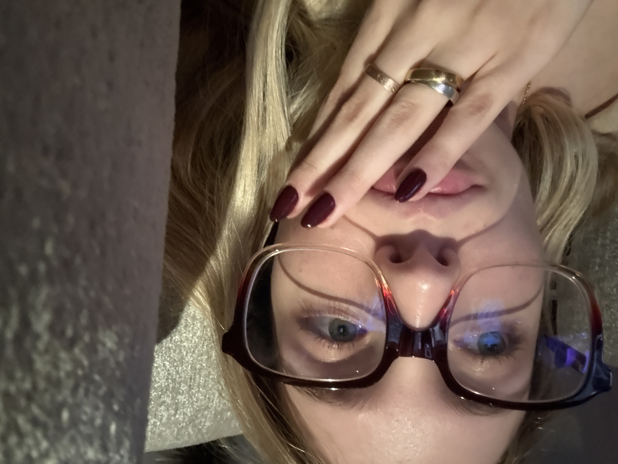 An upside-down close-up photograph depicts a fair-skinned person with blonde hair and blue eyes wearing red-framed glasses. Their right hand is positioned near their lips, revealing dark red nail polish and two rings. The person appears to be indoors, with a textured surface visible on the left side of the frame. The location within Mexico City is not discernible from the image.