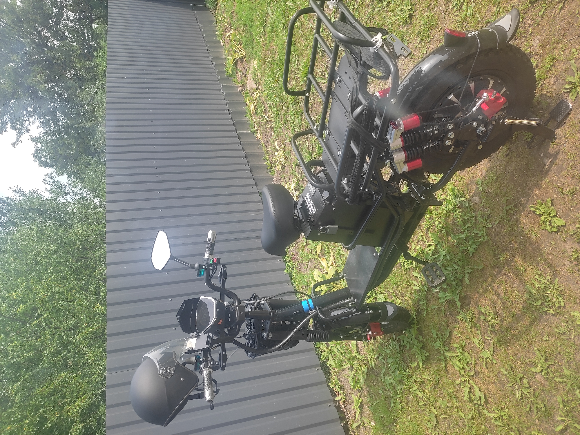 An electric scooter is parked on a grassy area in front of a grey corrugated metal fence. The scooter is black and has a helmet with a clear visor hanging from the handlebars. It also features a rear cargo rack, a black seat, and dual rear shock absorbers. The background consists of green trees. The ground is covered with grass and some dirt.