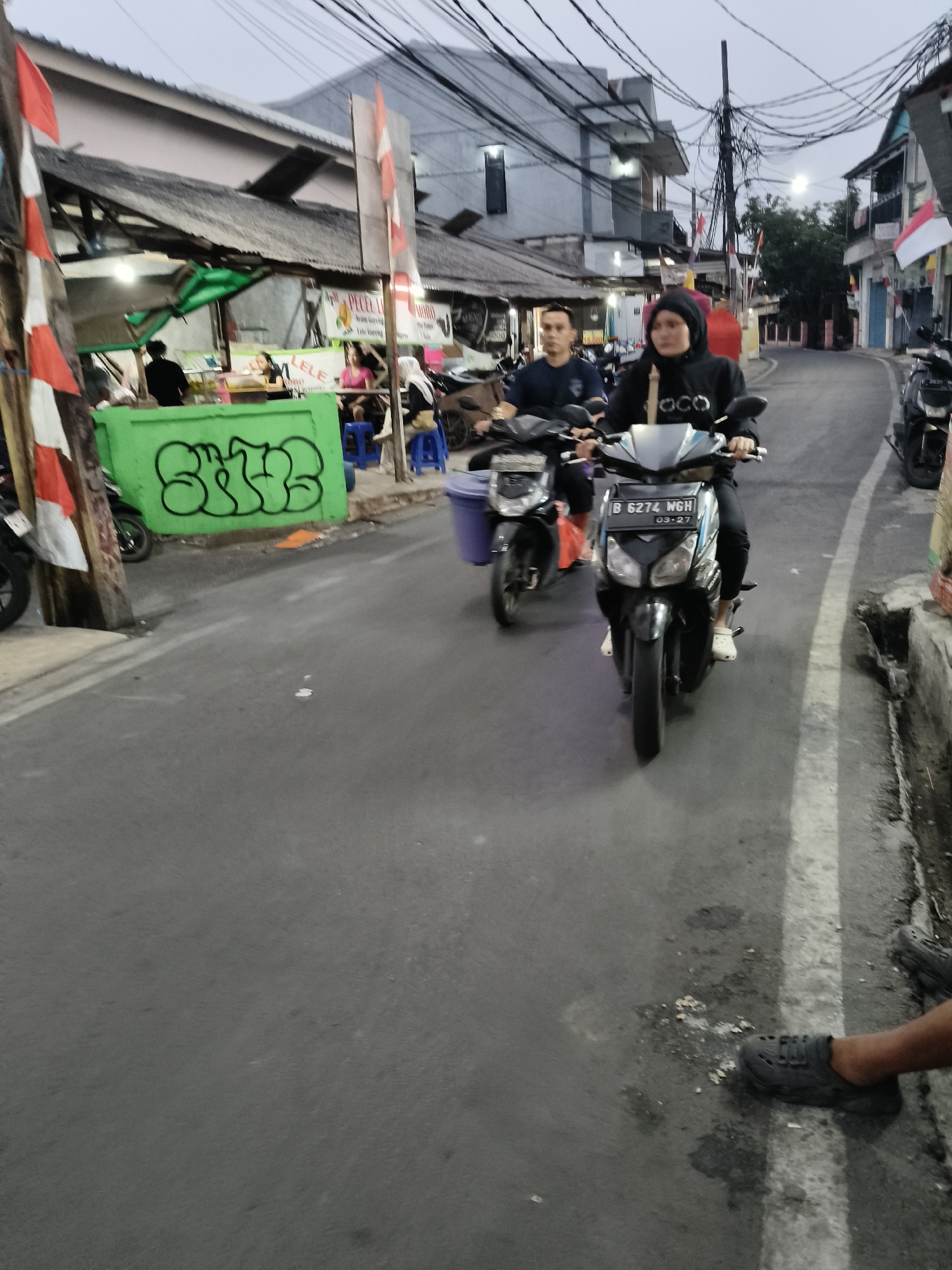 The image captures a street scene in South Tangerang, Indonesia, likely in the evening or early morning given the low light. Two individuals are riding motorcycles. The foreground shows the front of a motorcycle with the license plate "B 6274 WGH". The rider, wearing a black hijab and black hoodie, is looking forward, her face partially obscured by the hood. The second motorcycle, slightly behind and to the left, has a rider wearing a blue t-shirt and a passenger behind him.
The environment features a narrow street with buildings lining both sides. On the left, there are small shops with corrugated metal roofs and green and white banners, one with graffiti. Red and white Indonesian flags are visible on poles along the street. The road surface appears to be asphalt with some debris and puddles, suggesting it might be wet. Power lines are strung overhead, a common sight in urban areas. The overall impression is of a busy, local street in a residential or commercial area.