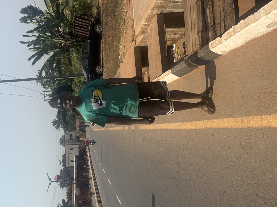 The image depicts a daytime scene on a street in Aguata, Nigeria. A dark-skinned male subject stands centrally in the foreground, facing the camera directly. He wears a black beanie, a green short-sleeved t-shirt with a "NIKE" logo, dark shorts with white side stripes and a white drawstring, and dark brown flip-flops. His hands are at his sides.
To the subject's left is an asphalt road with a solid yellow line delineating the shoulder and white dashed lines marking lanes further left. A person on a motorcycle is visible in the mid-ground, traveling away from the camera. In the background along the road, several buildings of varying sizes and designs are present, alongside modern solar streetlights.
To the subject's right, an unpaved dirt shoulder borders the road, followed by a concrete drainage ditch. The ditch is partially covered by concrete slabs and wooden planks, and contains visible refuse, including plastic items. Beyond the ditch and shoulder, a black sedan (consistent with a 1990s-2000s model Toyota Camry) is parked on a dirt patch, facing left. The vehicle's trunk appears open or damaged, with several metal frames, possibly window grilles, resting against it. Lush green foliage, including banana trees, densely covers the area behind the car and extends into the background. A wooden utility pole with overhead wires is situated near the sedan. The sky is clear, and direct sunlight illuminates the scene, casting shadows.