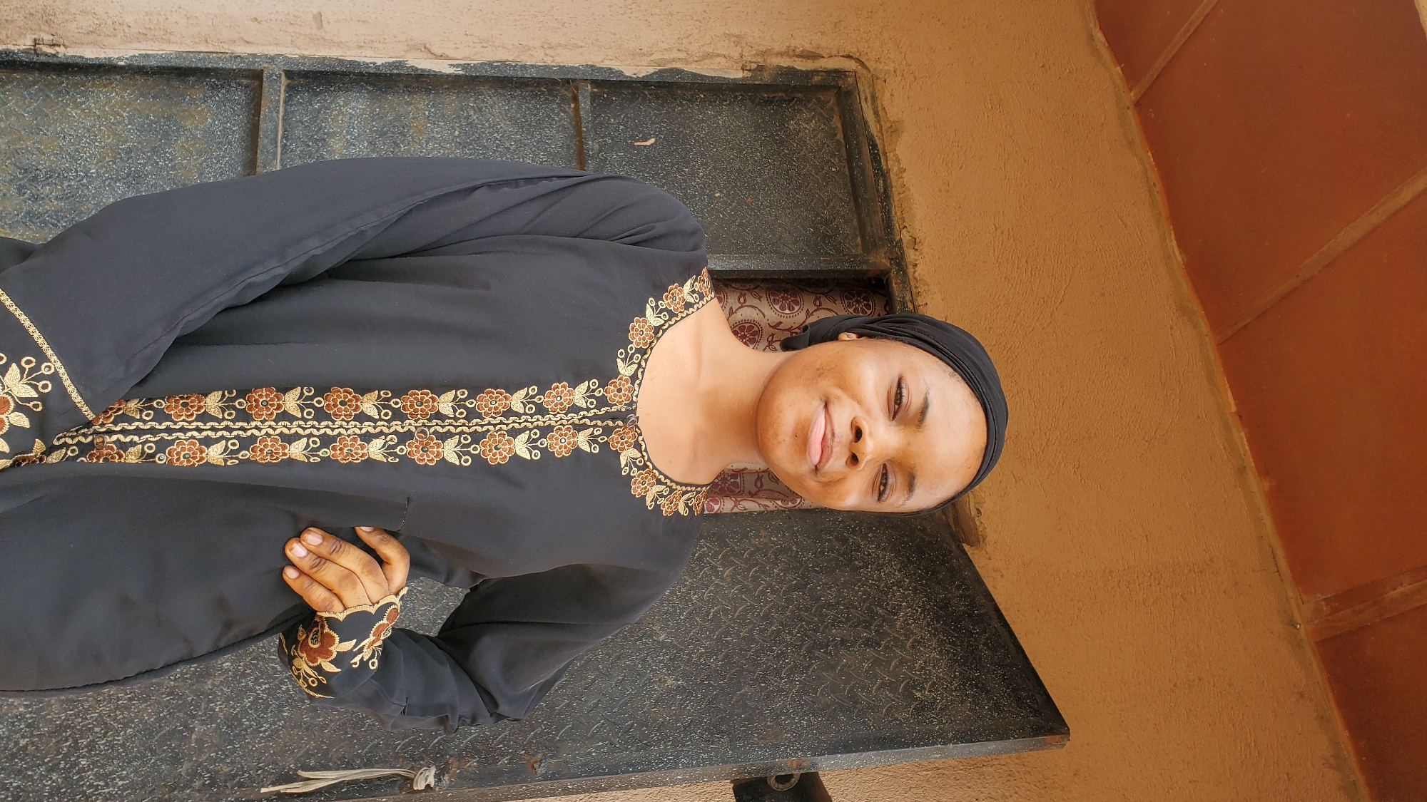 A young Black woman wearing a black hijab and a black abaya with gold and brown embroidery is standing in front of a building with a brown, textured wall and a dark metal door. She is smiling and looking at the camera, with her right hand resting on her hip. The door appears to be weathered and has some graffiti or dirt on it. The building is likely in an urban or semi-urban area, possibly in Jalingo, Nigeria, given the architectural style and materials commonly found in such locations. The lighting suggests it is daytime, possibly outdoors.