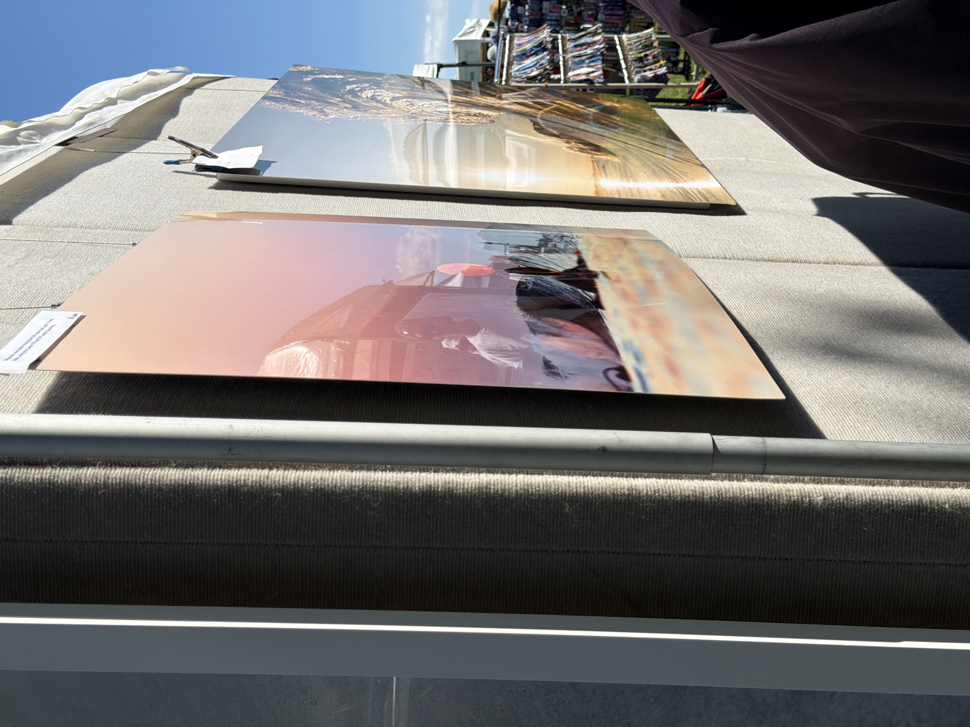 The image shows an outdoor art display, likely at a street fair or market, in the Village of Sag Harbor, United States of America. Two large, curved photographs are prominently displayed on a textured, light-colored wall. The photograph on the left features a warm-toned scene with a person in a white shirt standing in front of what appears to be a building or structure, with a pinkish hue cast over the image. The photograph on the right depicts a sunlit landscape, possibly a beach or water scene, with golden light reflecting on the surface. The photos are hung with thin wires, and one of them has a clothespin holding a small tag. In the background, to the right, a rack filled with colorful items, possibly merchandise, is visible. A person's shoulder and upper arm, wearing a dark garment, are in the foreground on the right side of the image, casting a shadow on the wall. The scene is illuminated by natural sunlight, creating strong contrasts and shadows. The sky above is a clear, bright blue.