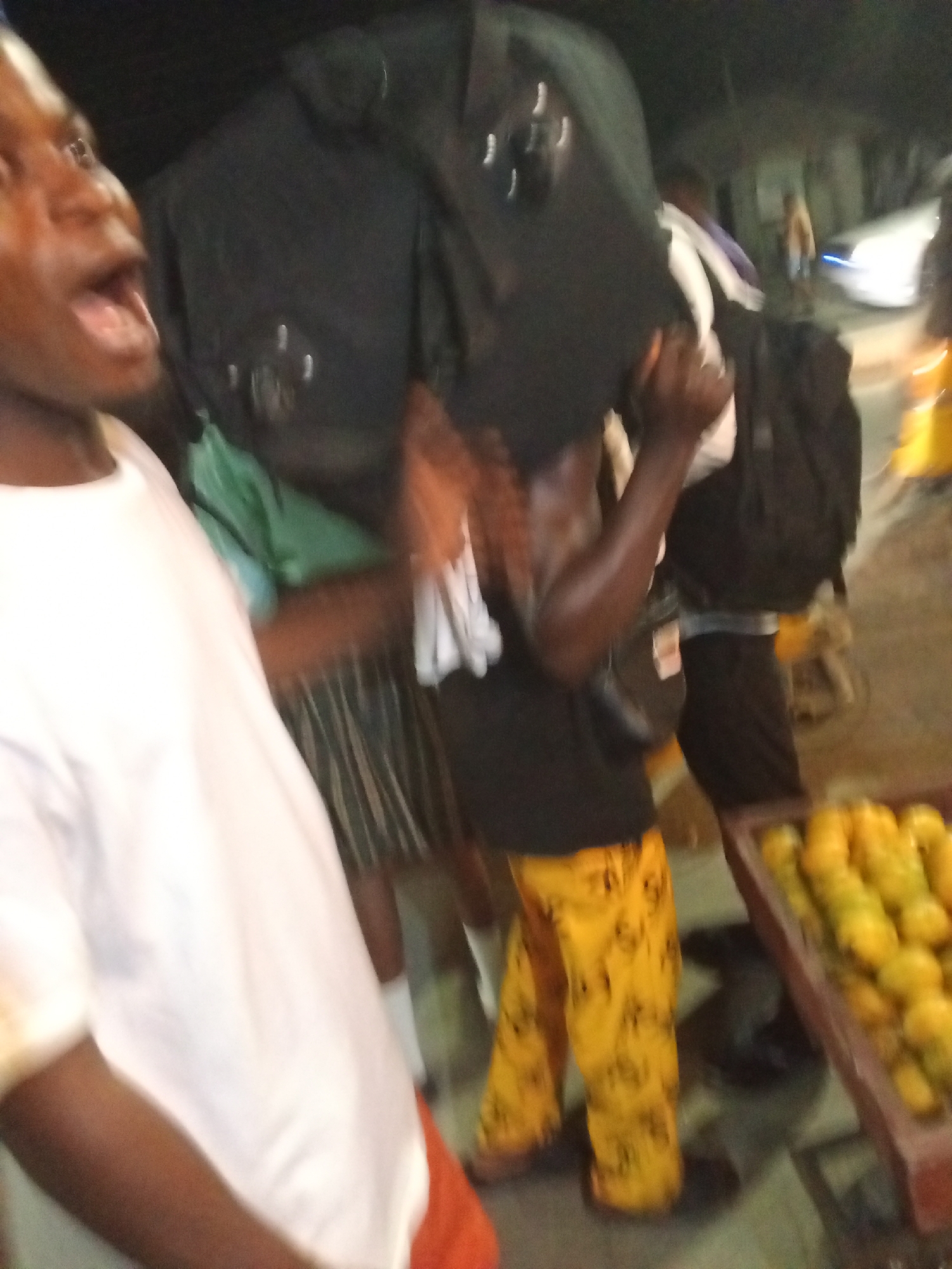 The image presents a blurry, low-light street or market scene, likely captured at night or dusk in Ikorodu, Nigeria. The overall impression is one of movement and activity in challenging lighting conditions.
In the immediate foreground on the left, a person, likely a young man with dark skin, is partially visible. He wears a white t-shirt, and his face is turned towards the right with his mouth wide open, suggesting he is speaking, shouting, or reacting to something.
Central to the composition, another dark-skinned individual is prominently featured, carrying a large, dark, bulky object or bag on their shoulder or head. This person wears a black t-shirt and distinctive yellow pants with a dark pattern. A white cloth is draped over their left shoulder, and their right arm is raised, possibly to stabilize the heavy load. This figure appears to be in motion.
To the right and slightly behind the central person, a third individual is partially visible, wearing a dark backpack. In the lower right corner of the frame, a brown or reddish crate is filled with numerous yellow or greenish-yellow fruits, strongly resembling mangoes, indicating a street vendor or market setting. In the blurred background, elements of a street are visible, including a white vehicle with illuminated headlights, further emphasizing the outdoor, night-time environment. No legible text is visible in the image.