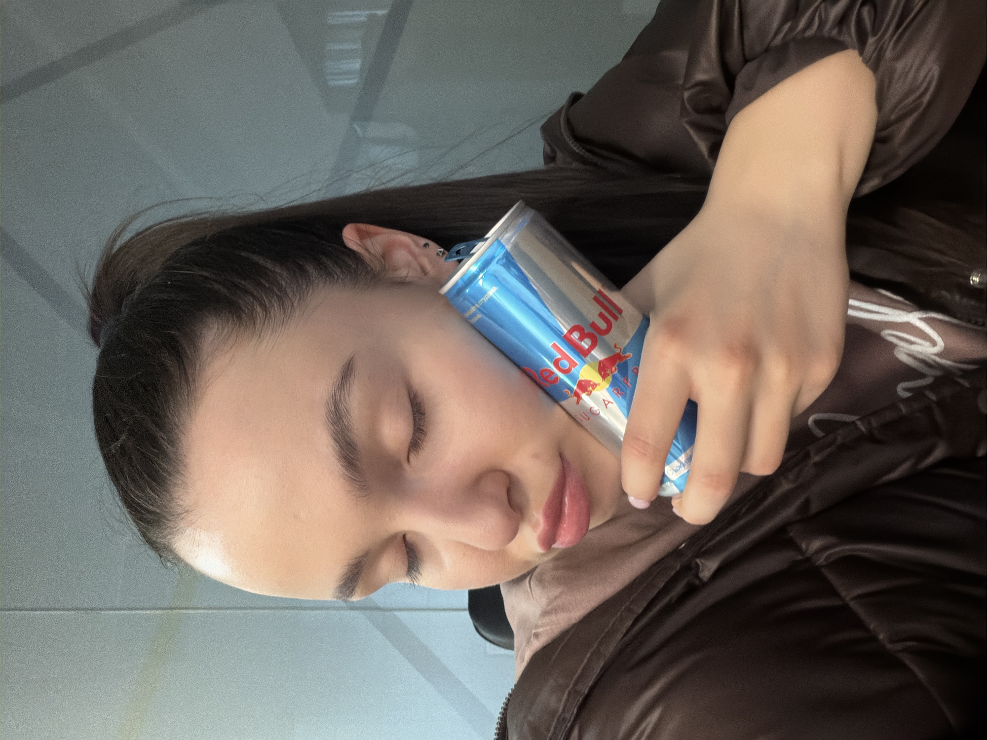 A young woman with dark hair is holding a blue and silver Red Bull energy drink can against her face. Her eyes are closed, and she appears to be resting. She is wearing a brown jacket over a light pink hooded sweatshirt. The background is a pale, possibly light gray or white, possibly a wall. Location is likely not discernible from this image alone. The city cannot be identified.