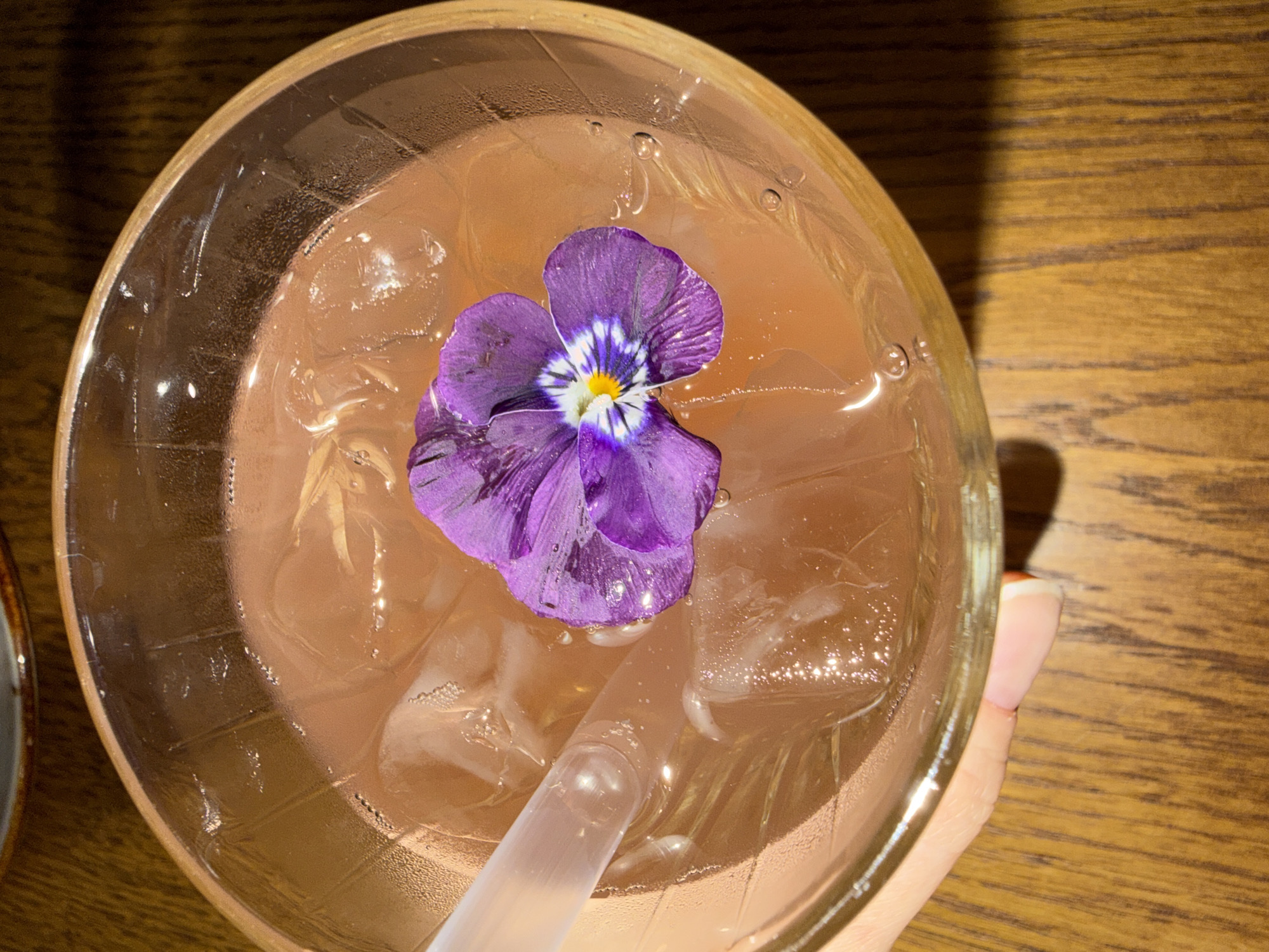 An overhead view captures a clear glass containing a light peach-colored liquid and multiple ice cubes. A single purple pansy flower with a yellow and white center floats on the liquid's surface. A translucent plastic straw is partially submerged in the drink, positioned to the left. Condensation droplets are visible on the exterior surface of the glass. A person's thumb and forefinger are partially visible on the lower left, gripping the glass. The glass rests on a wooden table exhibiting a distinct grain pattern. This scene is located in Khimki, Russia.