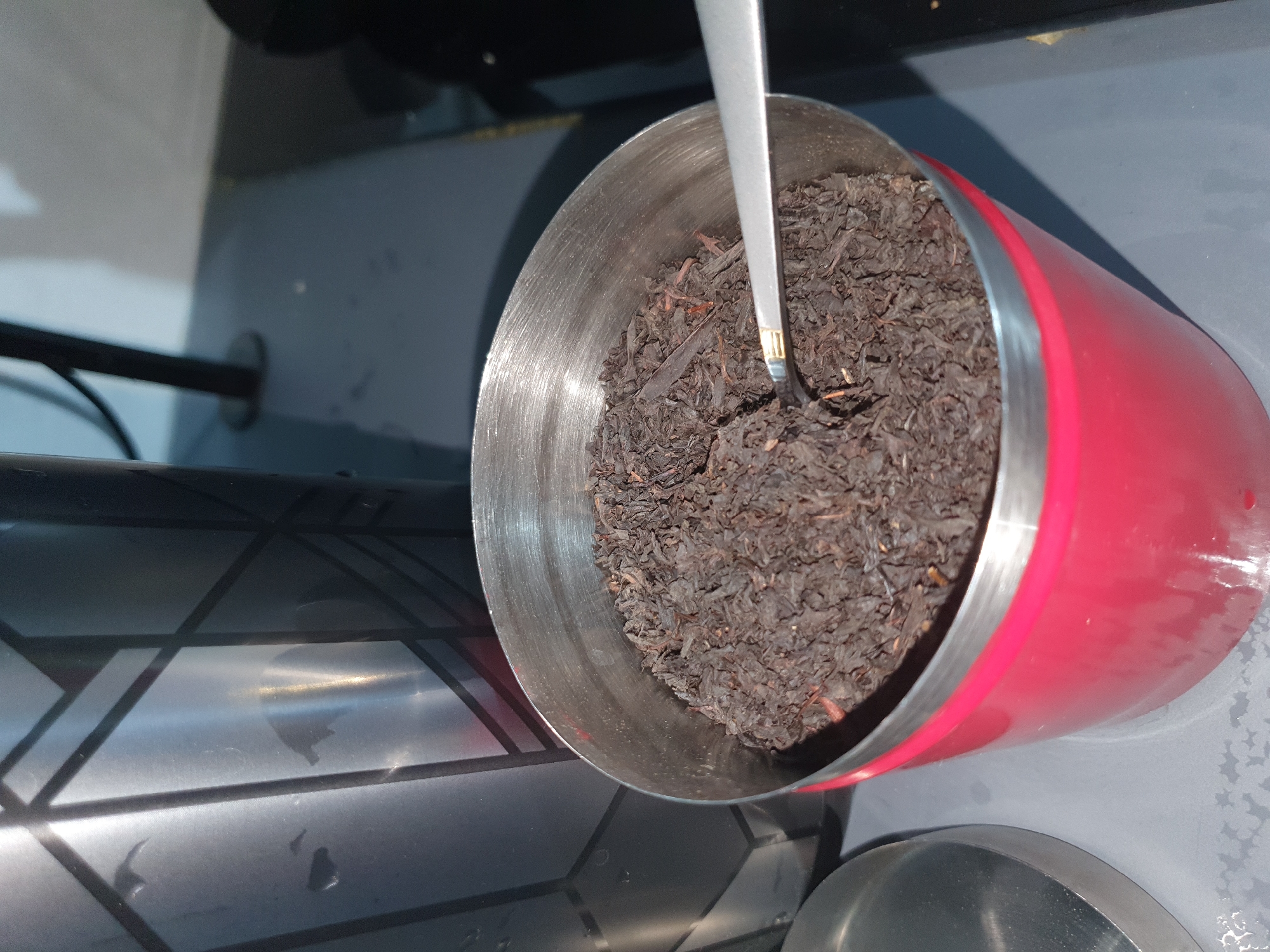 A close-up, eye-level shot shows a red metal container filled with loose-leaf black tea. A silver spoon is partially submerged in the tea. To the left of the red container is a silver lid, also appearing to be made of metal, and further to the left, a portion of a tall, silver thermos with a geometric pattern is visible. The objects are arranged on a grey surface, which may be a countertop or table. The background shows a blurred dark vertical object and a lighter grey area, possibly a wall or another surface. The lighting suggests an indoor environment, with a bright light source casting reflections on the metal objects. There are no people or animals visible in the image. The scene is situated within Kheyrabad, Iran.