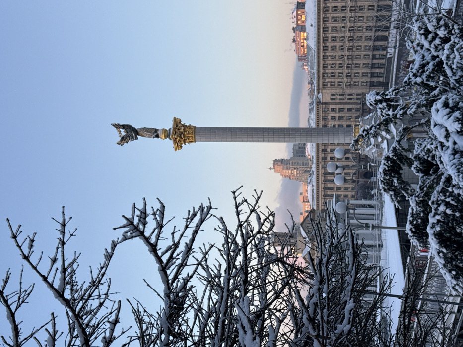 A statue of a winged figure atop a tall column stands prominently in the center. The city skyline rises in the background, with buildings visible behind the monument. The foreground features trees and shrubbery, their branches and leaves covered in snow. The sky is a light blue, suggesting a winter setting with the city partially dusted with snow.
