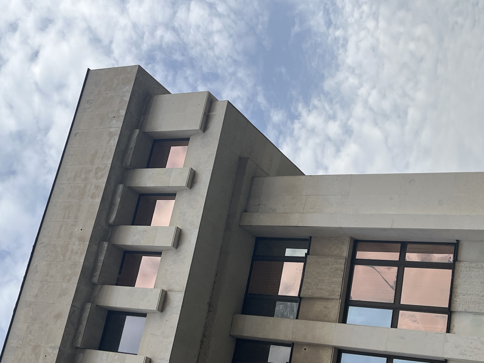 A low-angle shot captures a building in Burgas, Bulgaria, against a cloudy sky. The building features a light-colored exterior with a series of windows. The windows are reflective, mirroring the sky. The building's architecture includes projecting elements and a geometric design. The image suggests an urban environment, focusing on architectural details.