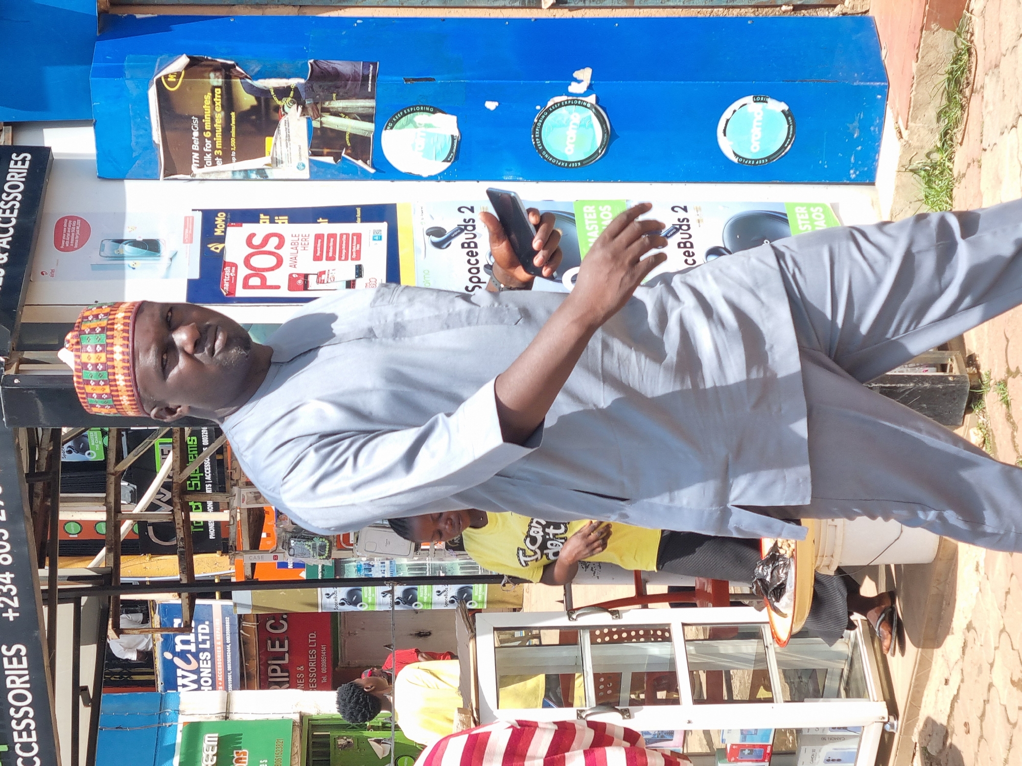 A medium shot captures a man in a gray traditional outfit and a colorful cap walking in an outdoor market environment. He holds a dark smartphone in his right hand, bringing it up towards his ear as if speaking or about to speak. The man is walking towards the viewer's right, with his body angled slightly to the left.
Behind him, a woman in a yellow t-shirt with the text "I Can Do It" is visible, also looking down. To the far left, a person in a striped red and white shirt is partially visible. The background is cluttered with various stalls and signage, suggesting a busy marketplace. One prominent sign displays "WIN PHONES LTD." and another has "TRIPLE C. ESSORIES LTD." The general setting appears to be a street in Jos, Nigeria, indicated by the signs with Nigerian phone numbers and company names common to the region. The pavement is cobblestone, and there is a visible drain. The lighting suggests it is daytime.