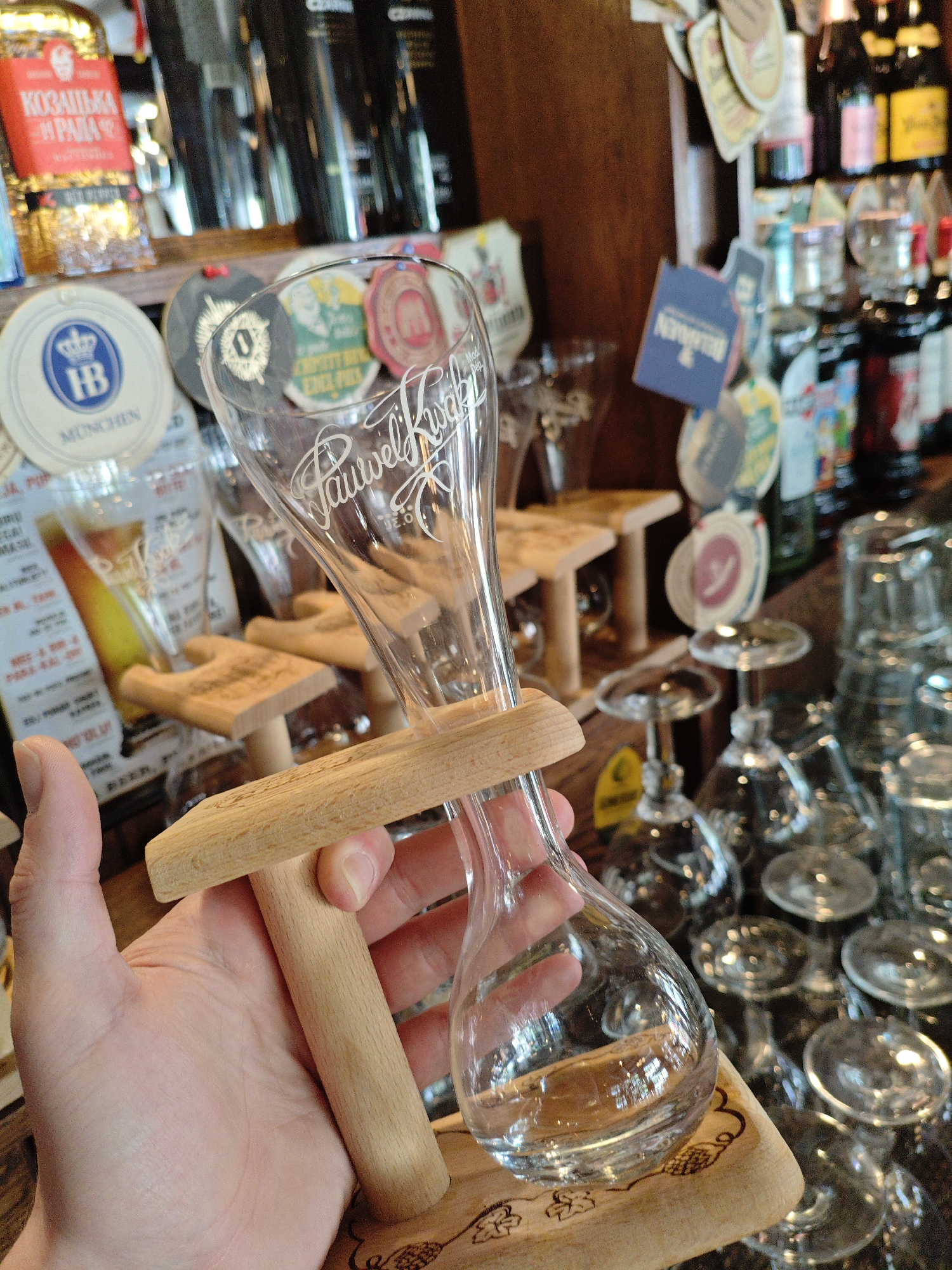A person's hand holds a clear glass beer mug with a wooden base. The mug is engraved with the name "Pauwel Kwak". The background shows a bar or pub interior with shelves stocked with bottles and glasses. Several beer coasters are visible on a shelf behind the mug, some featuring logos from German breweries. The scene appears to be in a bar or pub.