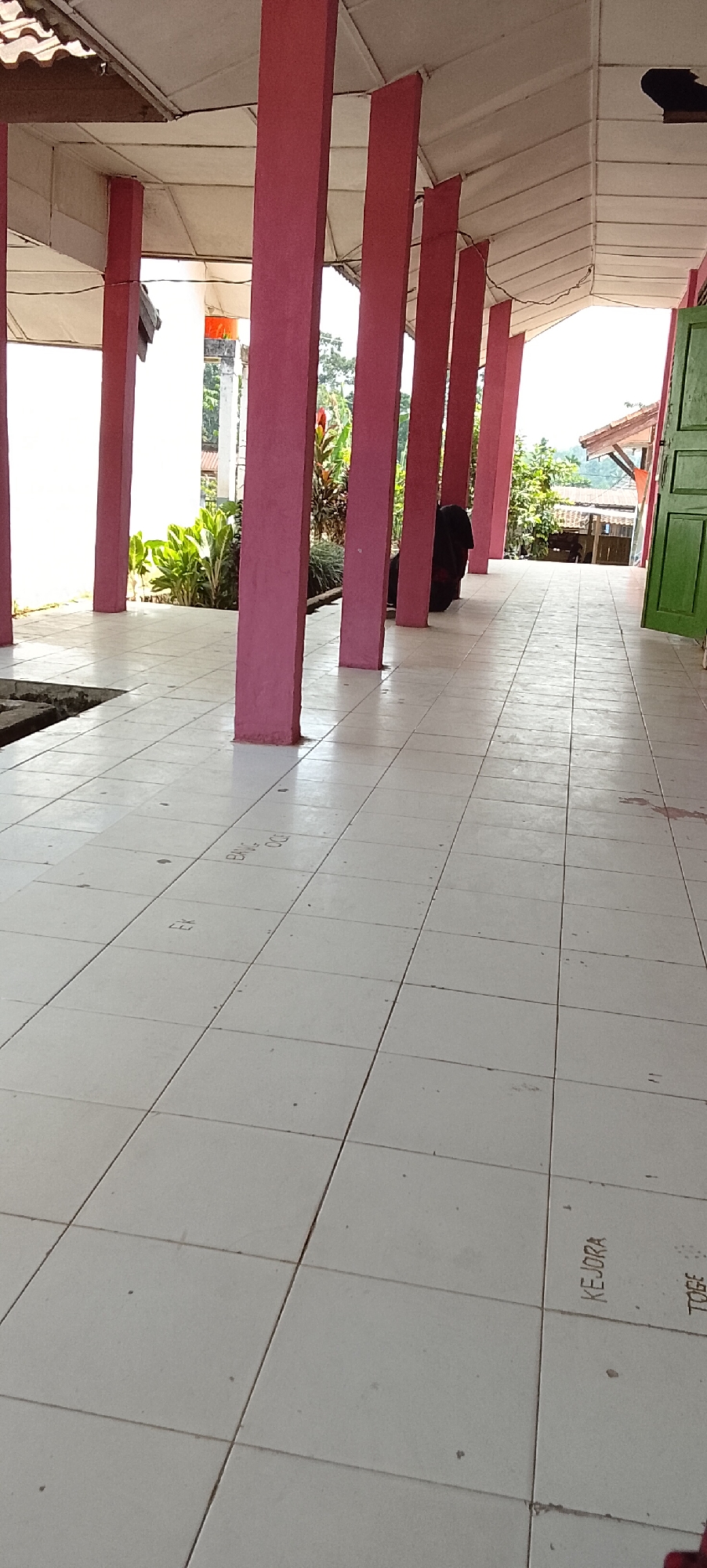 The image shows a covered walkway with a tiled floor in Jepara, Indonesia. Several large, reddish-pink pillars support the roof. Lush green foliage is visible behind the pillars on the left side of the frame, suggesting a tropical environment. A person, dressed in dark clothing, is seated on the ground behind one of the pillars, partially obscured from view. The tiled floor has some faint markings or writings on it. A green door is visible on the right side of the image, and further in the background, glimpses of buildings and trees indicate a residential or community area. The overall scene depicts an outdoor covered space, likely part of a building or facility in Jepara.