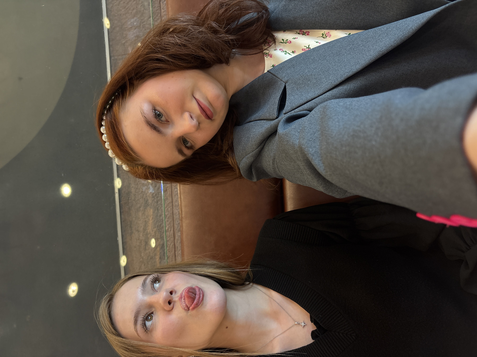 Two young women are captured in a selfie, likely taken indoors in Saint Petersburg, Russia. The image is rotated 90 degrees counter-clockwise.
The woman in the foreground on the right has long red hair, blue eyes, and is wearing a gray blazer over a white top with a subtle pink floral pattern. A pearl-like headband adorns her hair. She looks directly at the camera with a gentle, slightly smiling expression.
The woman in the background on the left has blonde hair and brown eyes. She is wearing a black long-sleeved top and a delicate silver necklace with a star-shaped pendant. She is making a playful face, sticking her tongue out and looking upwards and to the left of the frame.
The setting appears to be a modern interior space, possibly a cafe or lounge, with warm brown upholstered seating visible behind them. The background features dark, reflective surfaces that show circular light reflections, suggesting overhead lighting. No text is visible in the image.