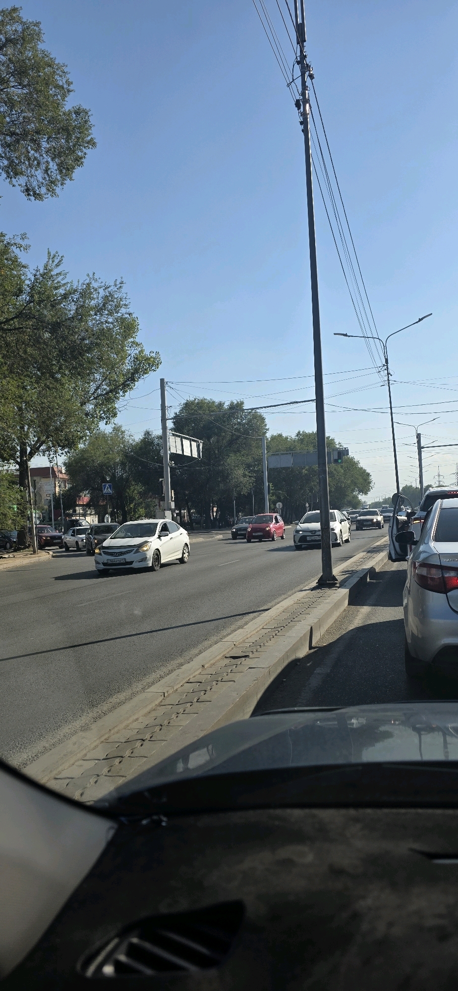 The image depicts a street scene in Almaty, Kazakhstan, during daylight. The perspective is from within a vehicle, showing the road ahead. Several cars are visible, including a white sedan in the foreground on the left, a red sedan further down the road, and a silver sedan on the right. There are traffic lights, streetlights, and utility poles with power lines crisscrossing the sky. Trees line the side of the road, and buildings are partially visible in the background. The sky is clear and blue. The road appears to be a multi-lane thoroughfare with a sidewalk and curb. The overall impression is of a typical urban traffic environment on a sunny day.