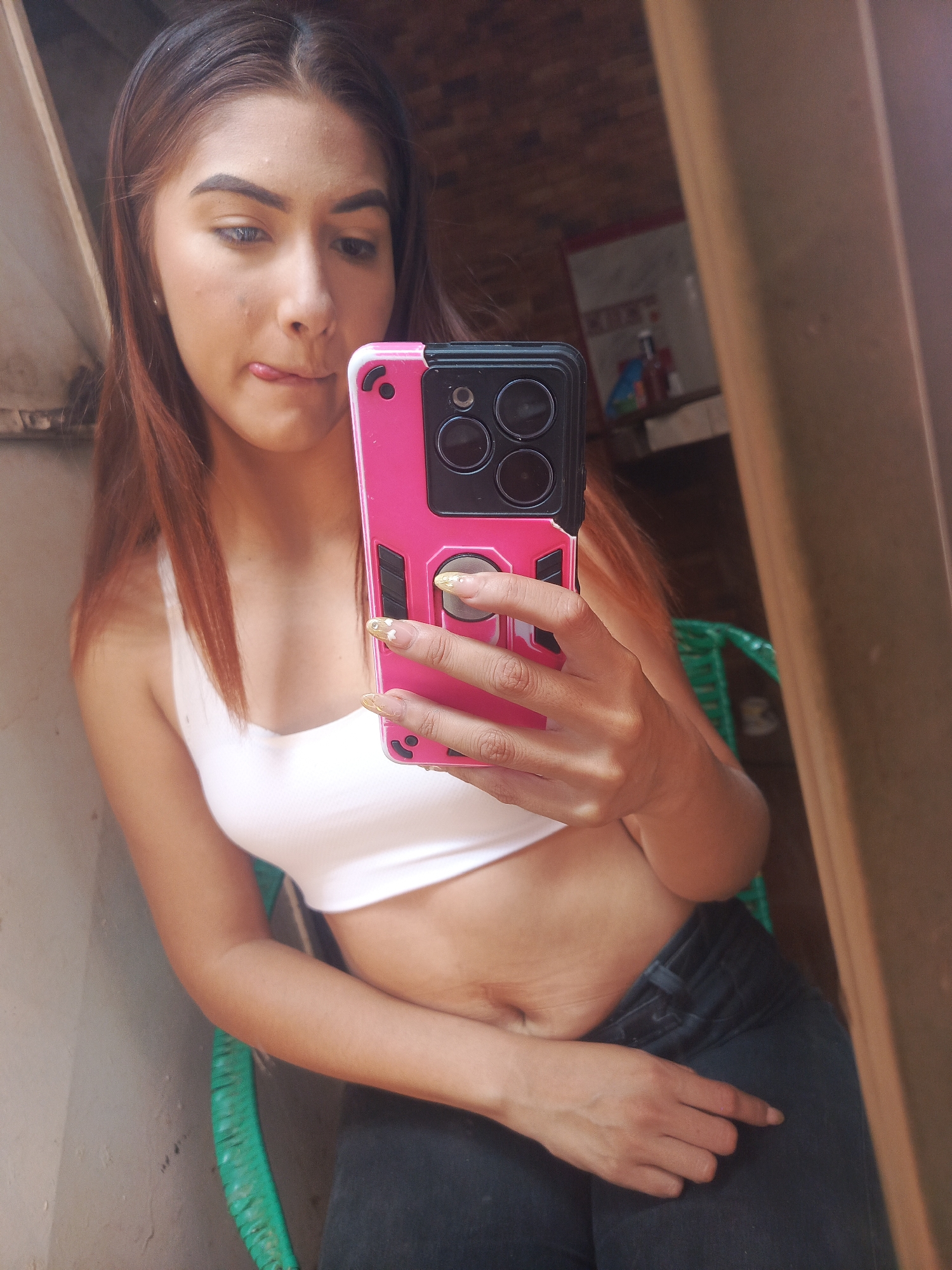 A young woman with reddish-brown hair is taking a selfie in what appears to be an interior space. She is wearing a white crop top and dark denim jeans. She is holding a smartphone with a pink protective case, covering part of her face and torso. The woman has her tongue slightly out in a playful expression. In the blurred background, there is a green plastic chair and what seems to be a shelf with various objects. The location is identified as Juan Griego, Venezuela.