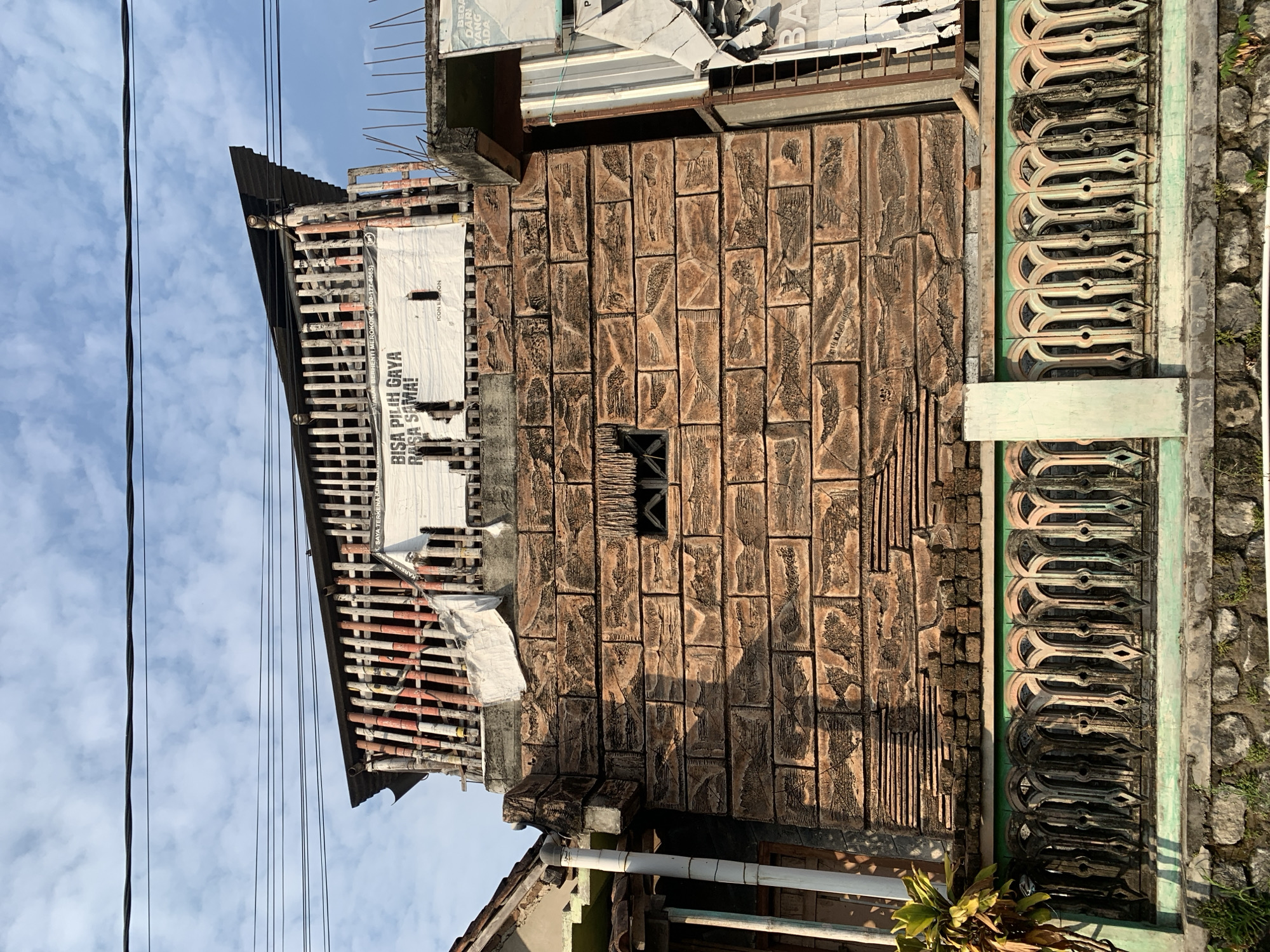 The image shows a portion of a building with a textured brick facade, located in Tanjungsepreh, Indonesia. Above the brick wall, there is a structure with vertical wooden slats, some painted red and white. A white banner with Indonesian text, "BISA PILIH GAYA RASA SAMA!", is attached to this structure. There are also power lines running across the top of the frame against a blue sky with clouds. A decorative green and cream colored railing runs along the top of a stone wall in the foreground. To the right of the main building, a section of another building is visible, with torn posters on its facade. The overall scene suggests a partially constructed or aged urban environment.
