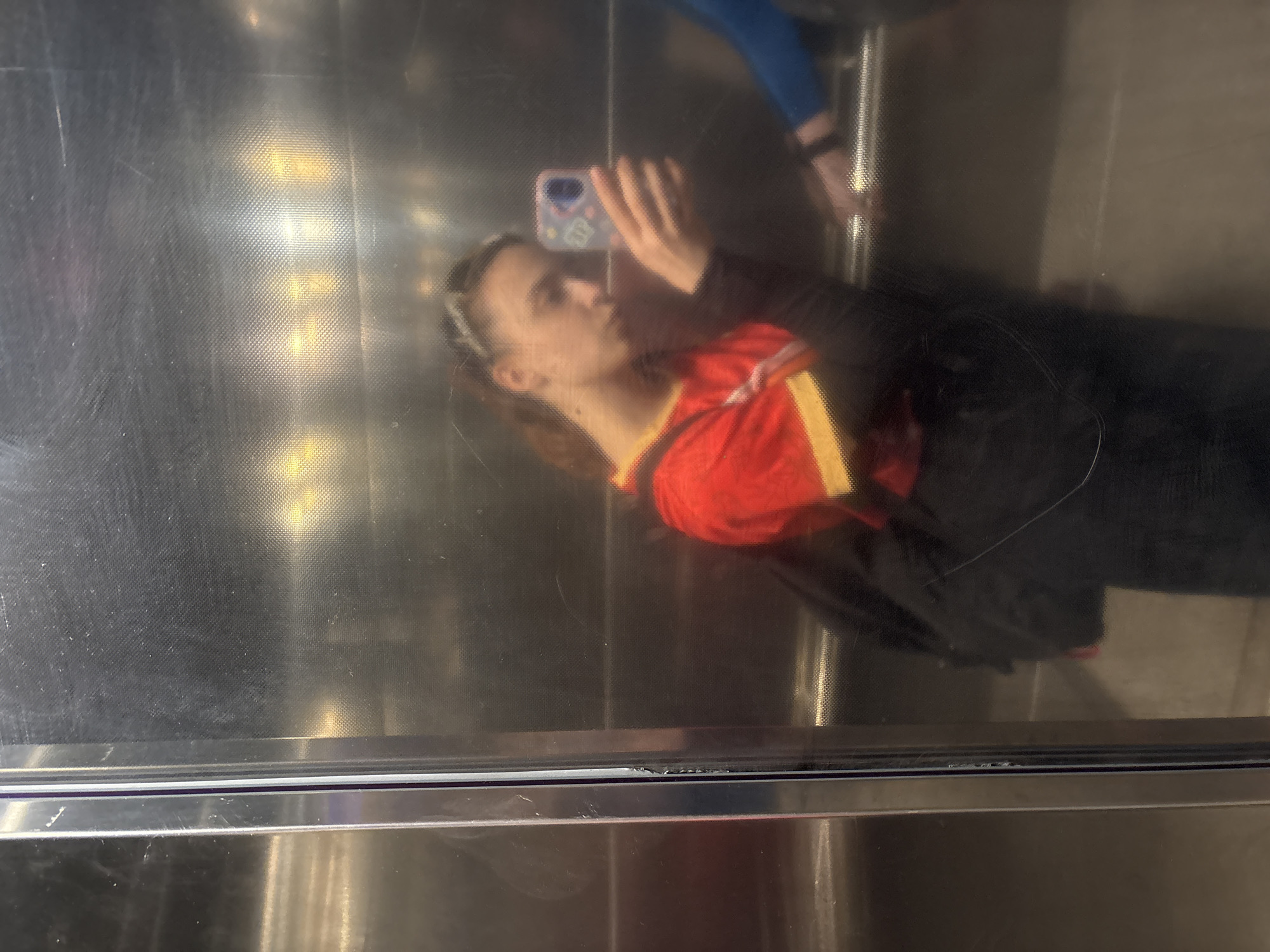 A person is taking a selfie in what appears to be an elevator. The individual is wearing a red shirt with a gold stripe and a black jacket. They are holding a smartphone in their right hand. The elevator has metal surfaces. It is unclear if the city of Perm, Russia, is related to the image's location.