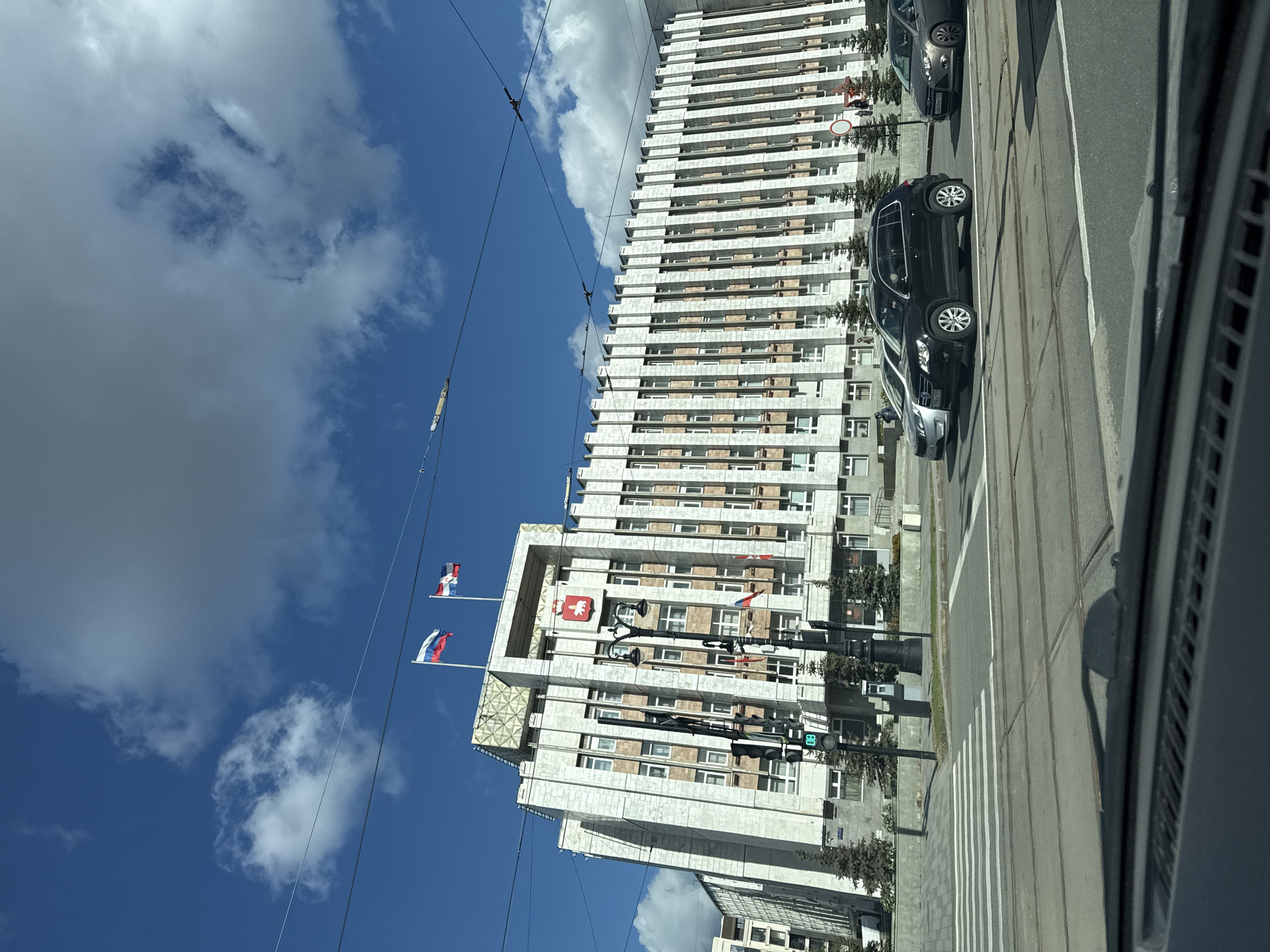 The image captures a view of the city of Perm, Russia, specifically focusing on the building housing the Legislative Assembly of Perm Krai, easily recognizable by the flags displayed on its roof. The architectural style is a mix of Soviet-era modernism with some decorative elements. Several vehicles are parked along the roadside. The sky above is blue with scattered white clouds. Overhead power lines are visible. The image appears to be taken from a moving vehicle.