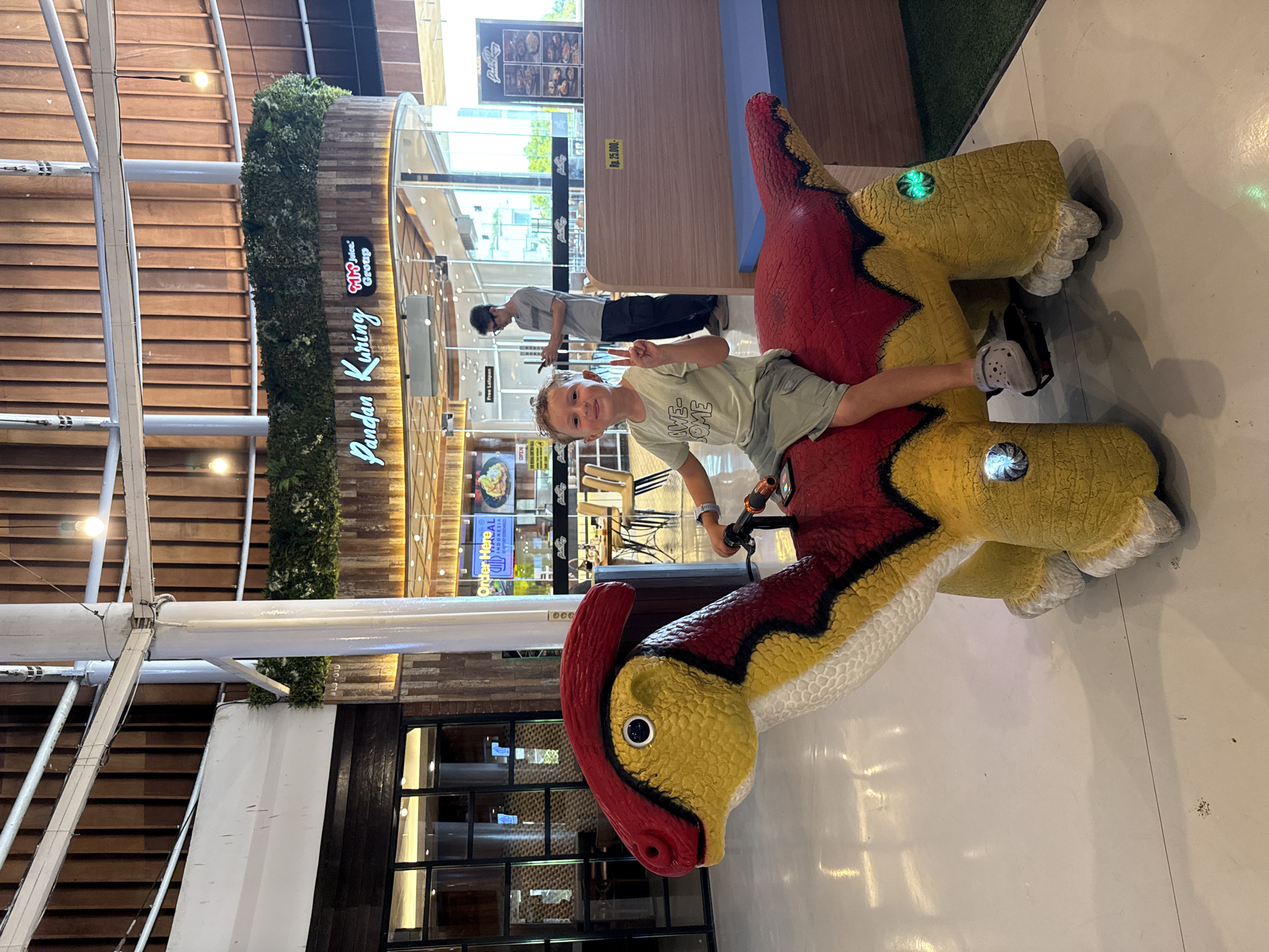 In Kuta, Indonesia, a young boy, approximately 7 years old, is seated on a yellow and red dinosaur-shaped ride-on toy in a well-lit, indoor commercial space. The boy is wearing a light green t-shirt with the text "AWESOME" and "SOME" visible, and light gray shorts. He has short, light brown hair and is making a peace sign with his right hand while smiling at the camera. The dinosaur toy is brightly colored with red accents on its head and back, and yellow body with black detailing. It has blinking green lights on its sides. The boy's feet are resting on black pedals. In the background, a man wearing a gray t-shirt and dark cargo pants is standing and looking at his phone. Beyond him, there are tables and chairs, suggesting a dining area. Above, a curved ceiling with wooden paneling is visible, along with hanging light fixtures. A sign with the text "Pandan Kuring" is illuminated in the background, indicating the name of a restaurant. The floor is a light-colored polished surface. The overall environment suggests a family-friendly entertainment or dining complex.