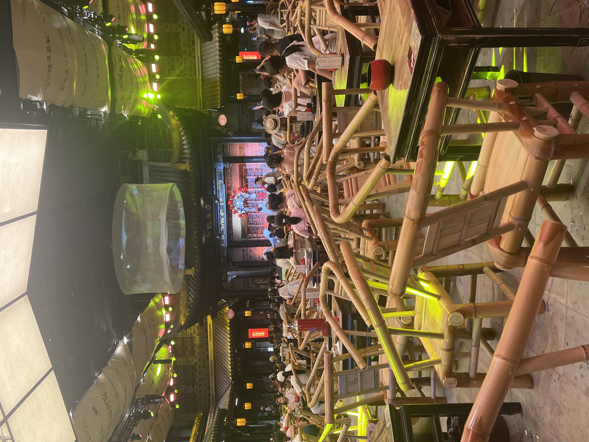 The image shows an indoor seating area with many bamboo chairs and tables. The area appears to be a restaurant or entertainment venue, with a stage set up at the far end of the room where a performance is taking place. Several people are seated at the tables, watching the performance or interacting with each other. The lighting is dim, with some spotlights directed towards the stage and the seating area. The architecture of the building suggests a traditional Chinese style, with ornate roof designs and lanterns. The overall atmosphere is lively and social, with people enjoying a night out.
