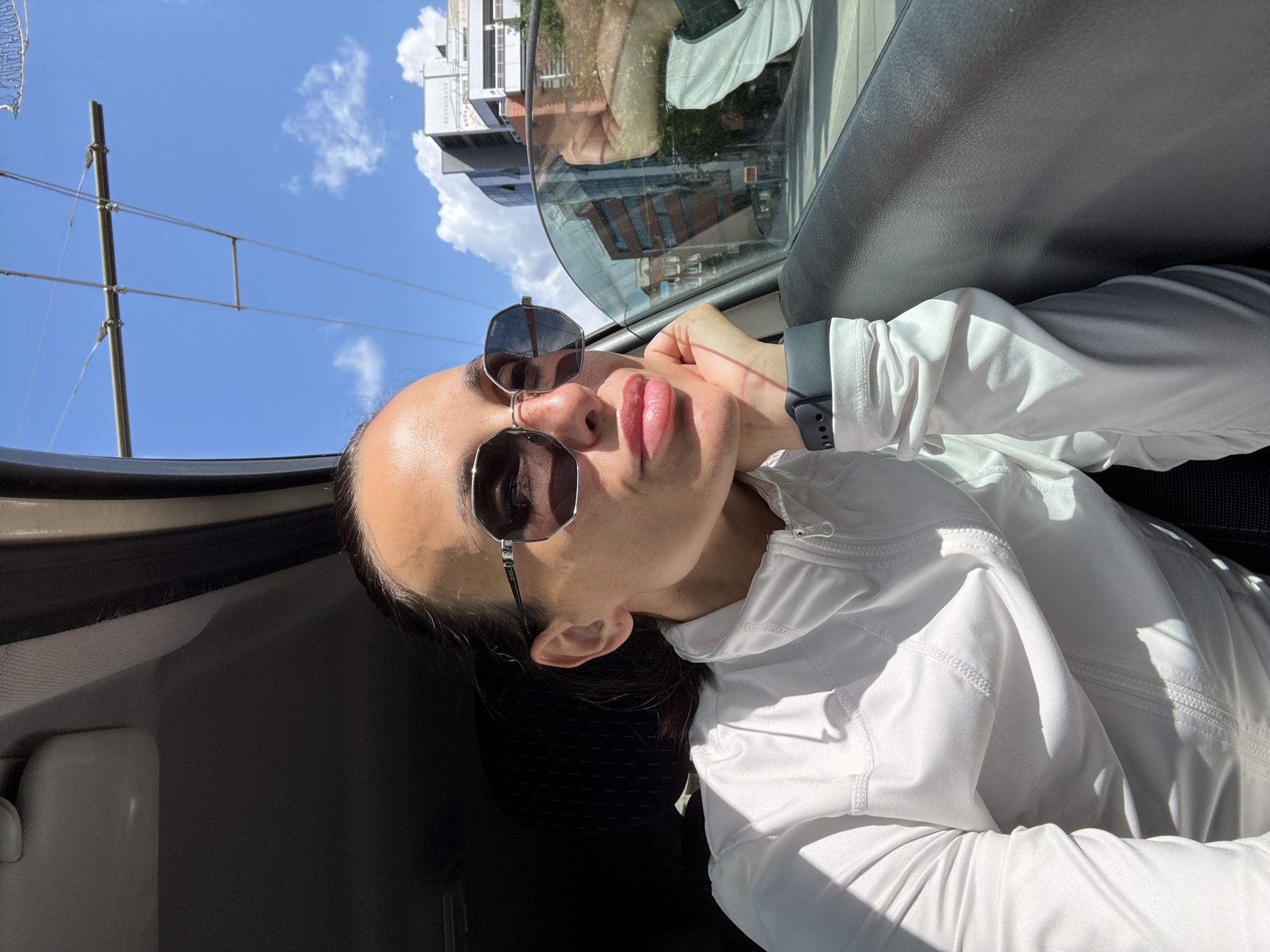 A woman wearing sunglasses and a white zippered jacket is in the passenger seat of a car, looking out the window. Her hand is resting on her chin. The sky is blue with scattered clouds. In the background, buildings and power lines are visible. The car appears to be in an urban area with trolley tracks. The scene is lit by bright sunlight, casting shadows on the woman and the interior of the car. The location is identified as Pleven, Bulgaria.