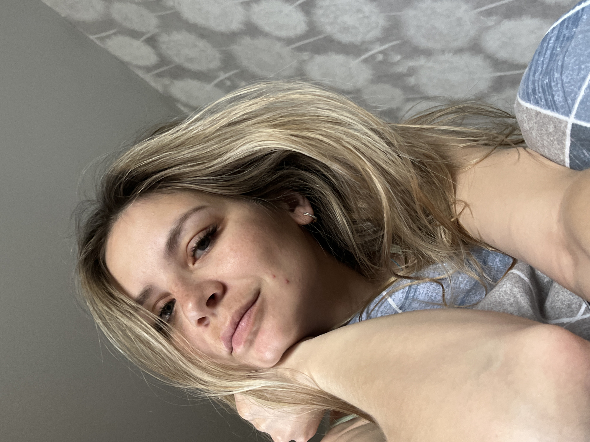 A woman with blond hair is lying down. She is resting her face on her arm. The background consists of a wall with a patterned design. There is no indication of the location being in Chaikovsky, Russia.