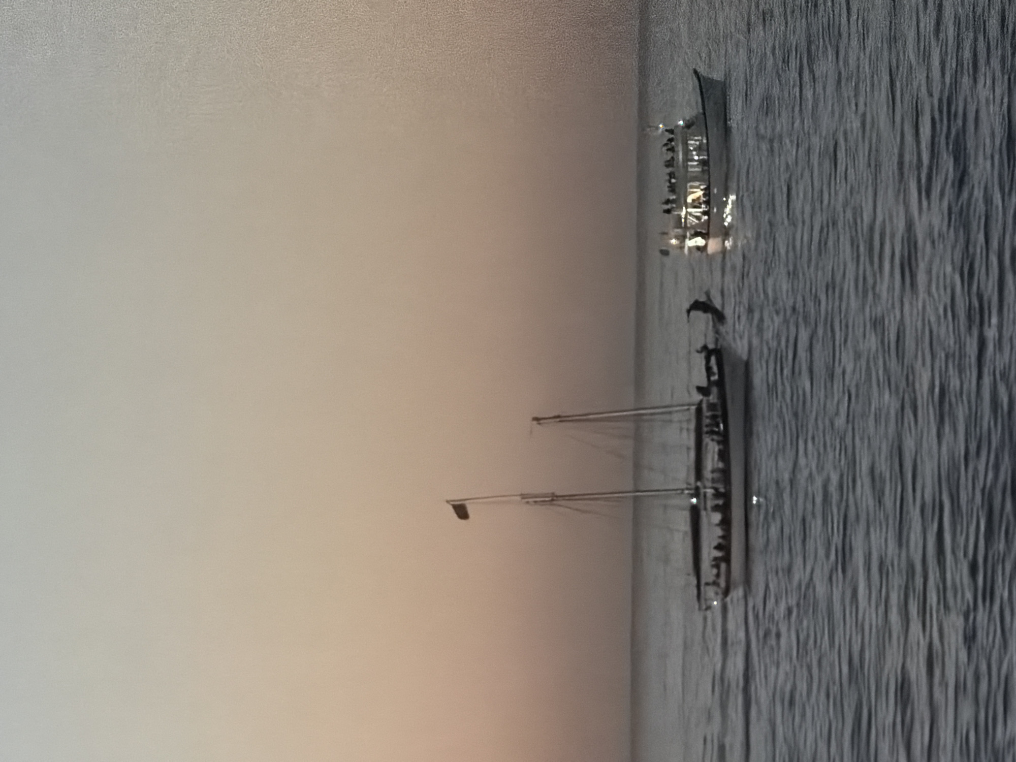 The image depicts two boats on the water at sunset or sunrise. The sky is light with a gentle gradient. The water appears choppy. One boat is a large sailboat with two visible masts and people onboard. The other boat is smaller and illuminated with lights, also with people onboard. Location is indiscernible.
