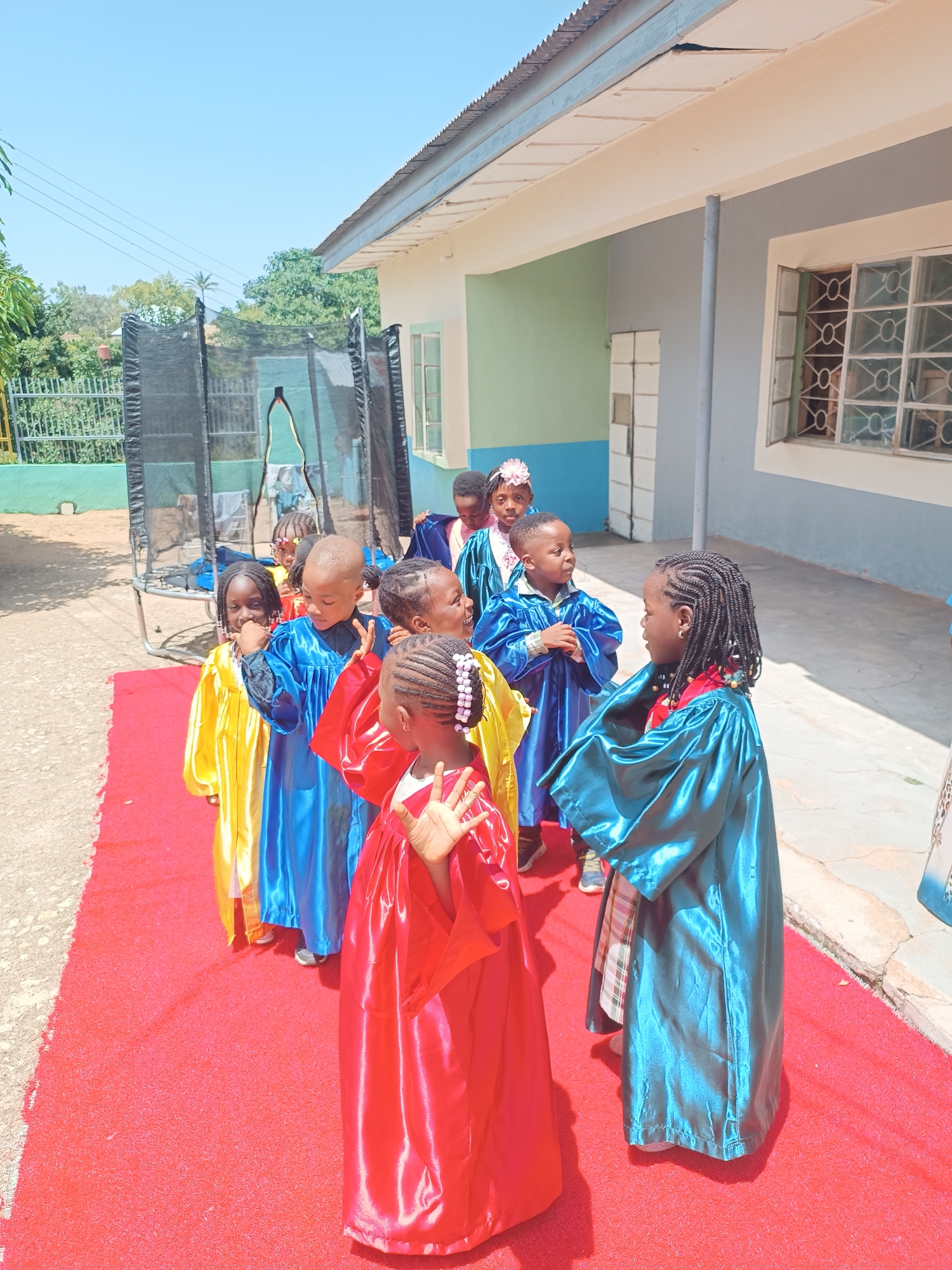 A group of young children, likely students, are gathered outdoors on a sunny day, standing on a red carpet in front of a building. They are dressed in colorful academic gowns, predominantly in shades of yellow, blue, and red, with some wearing matching caps or headbands. Their hair is styled in various braids, and some have decorative beads. The building appears to be a school, with a light blue and grey exterior, featuring a corrugated metal roof and windows with decorative grilles. A trampoline is visible in the background to the left, suggesting a playground area. The environment appears to be an urban setting, consistent with locations in Jos, Nigeria, where such educational institutions and outdoor spaces are common. The children are looking in various directions, some interacting with each other, while others appear to be posing or waiting. The scene captures a moment of celebration or a formal school event.