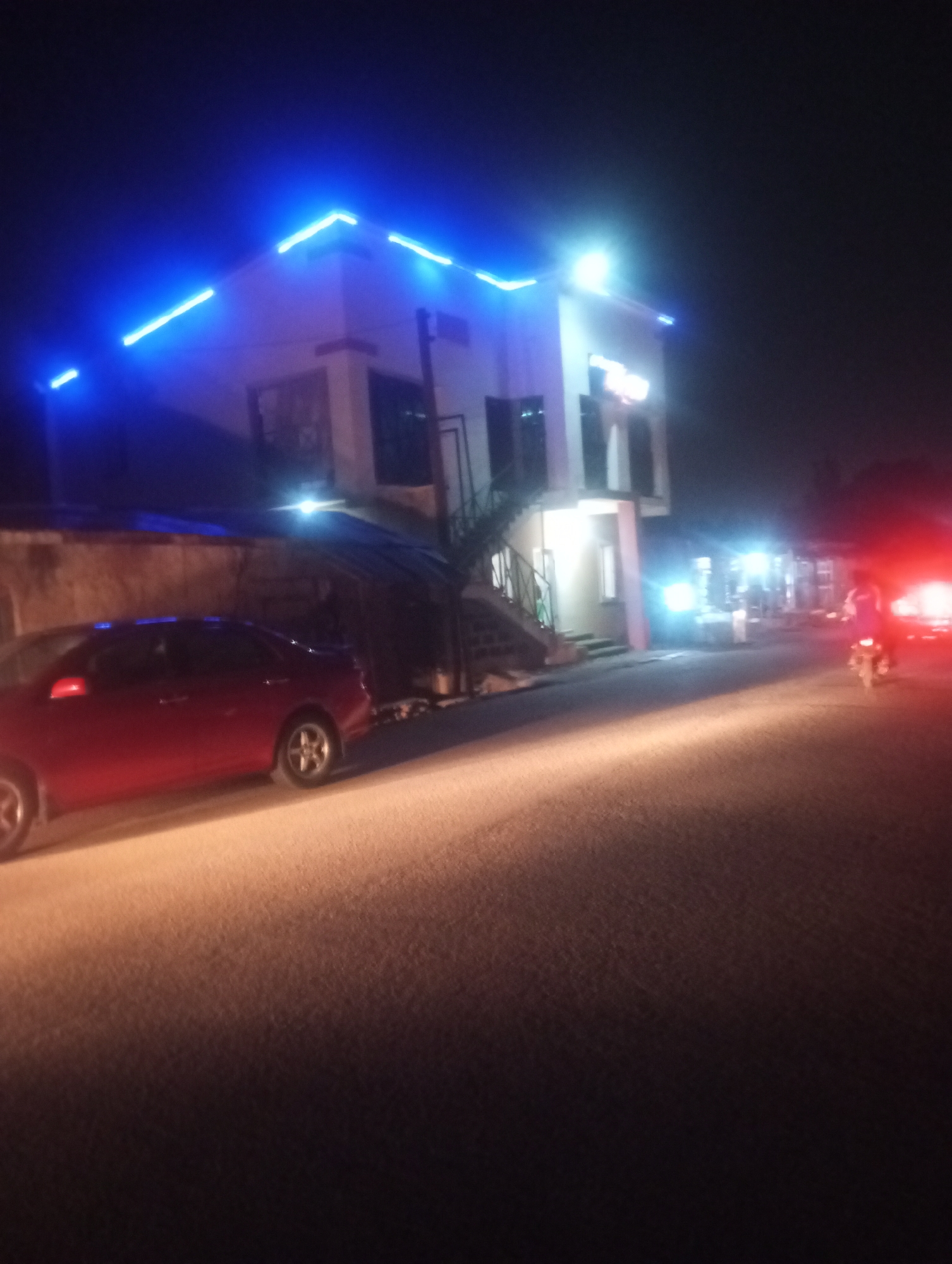 The image captures a night scene on a street, likely in Jos, Nigeria. The time of day is clearly night, characterized by the dark sky and reliance on artificial illumination.
The main subject is a two-story building located on the left side of the road. Its roofline is strikingly adorned with bright blue LED strip lights, creating a modern and eye-catching glow. The building also features several white lights on its facade and interior, with a prominent bright light source on its upper right section. Steps lead up to an entrance on the left side of the building.
In front of the building, a red sedan is parked along the roadside. The car's side is illuminated by a strong, diagonal beam of light that cuts across the paved road, creating a dramatic highlight. Further down the street on the right, a motorcycle with a rider is seen moving away from the viewer, its bright red taillight visible. Other bright white lights are scattered further down the street on the right, suggesting more establishments or activity.
The road surface is visible, brightly lit in sections by the aforementioned light beam and the motorcycle's lights, contrasting with the darker areas. No discernible text is visible on any signs or objects within the frame. The overall impression is a bustling street scene after dark, illuminated by various artificial light sources.