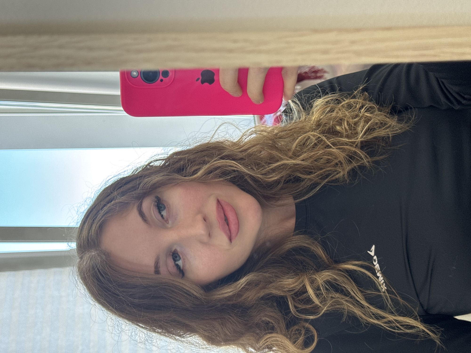 A woman with long, wavy blonde hair is taking a selfie with a pink iPhone in front of a window with blinds. She is wearing a black long-sleeved athletic shirt with the word "DEMIX" in white. The location is likely in Chaikovsky, Russia, but this cannot be definitively determined from the image.