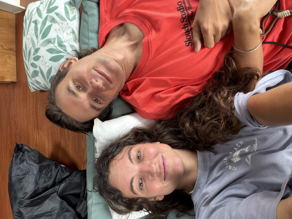 Two individuals lie on their backs, resting their heads on pillows. One wears a red shirt with "Slouch Studio Wine Napa" printed on it. The other person is in a grey shirt, and some text printed that is not readable. They both look up at the camera. A dark bag sits on the floor next to them. There are some bedsheets and the pillow has a green pattern. The floor has some wood.