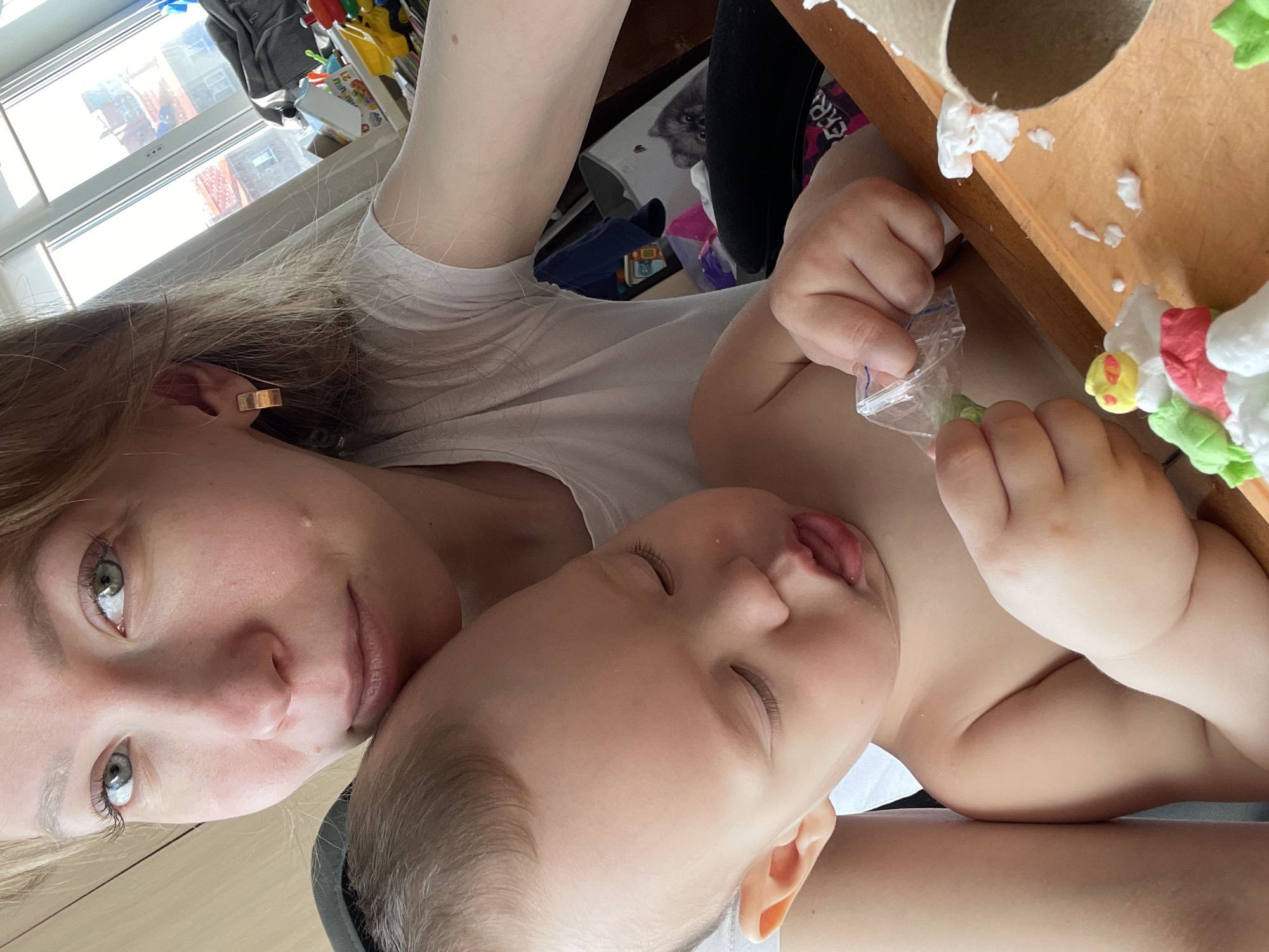 A selfie image shows a woman with fair skin, green eyes, and blond hair wearing a white T-shirt. She is facing the camera with a baby lying on her chest, possibly asleep. The baby is fair-skinned with closed eyes.
To the right of the baby, there's a table with a cardboard tube, small pieces of white material, and a decorated object with green, red, yellow, and white parts. The baby's hand is holding a clear plastic bag.
In the background, a window shows a cityscape, and there are various toys and objects, including a gray backpack. The scene appears to be indoors, potentially in a home environment. The location in Perm, Russia, is not verifiable from the image.