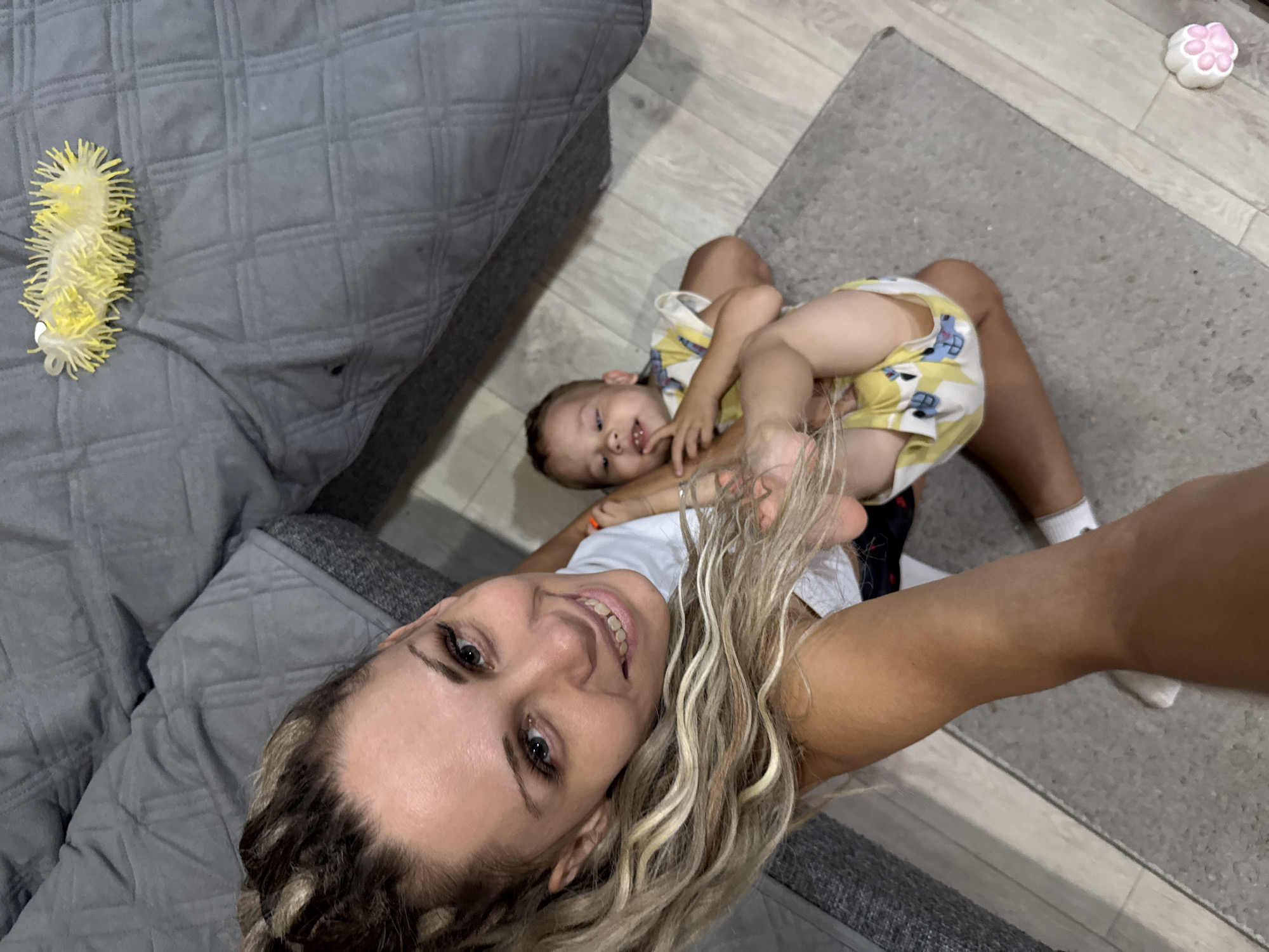 A high-angle, eye-level, full body shot shows a woman and a young child on a gray rug in what appears to be a living room. The woman, with blonde hair styled in a high ponytail, smiles at the camera and is wearing a white t-shirt. She is sitting on the rug, with her legs extended toward the camera. The child, who is wearing a yellow and white outfit with a cartoon print, is sitting in front of the woman, facing away from the camera. The child's left arm is extended towards the woman. In the background, a large gray upholstered couch is visible. A yellow, spiky toy is placed on the couch. The floor is made of light-colored wood planks.
