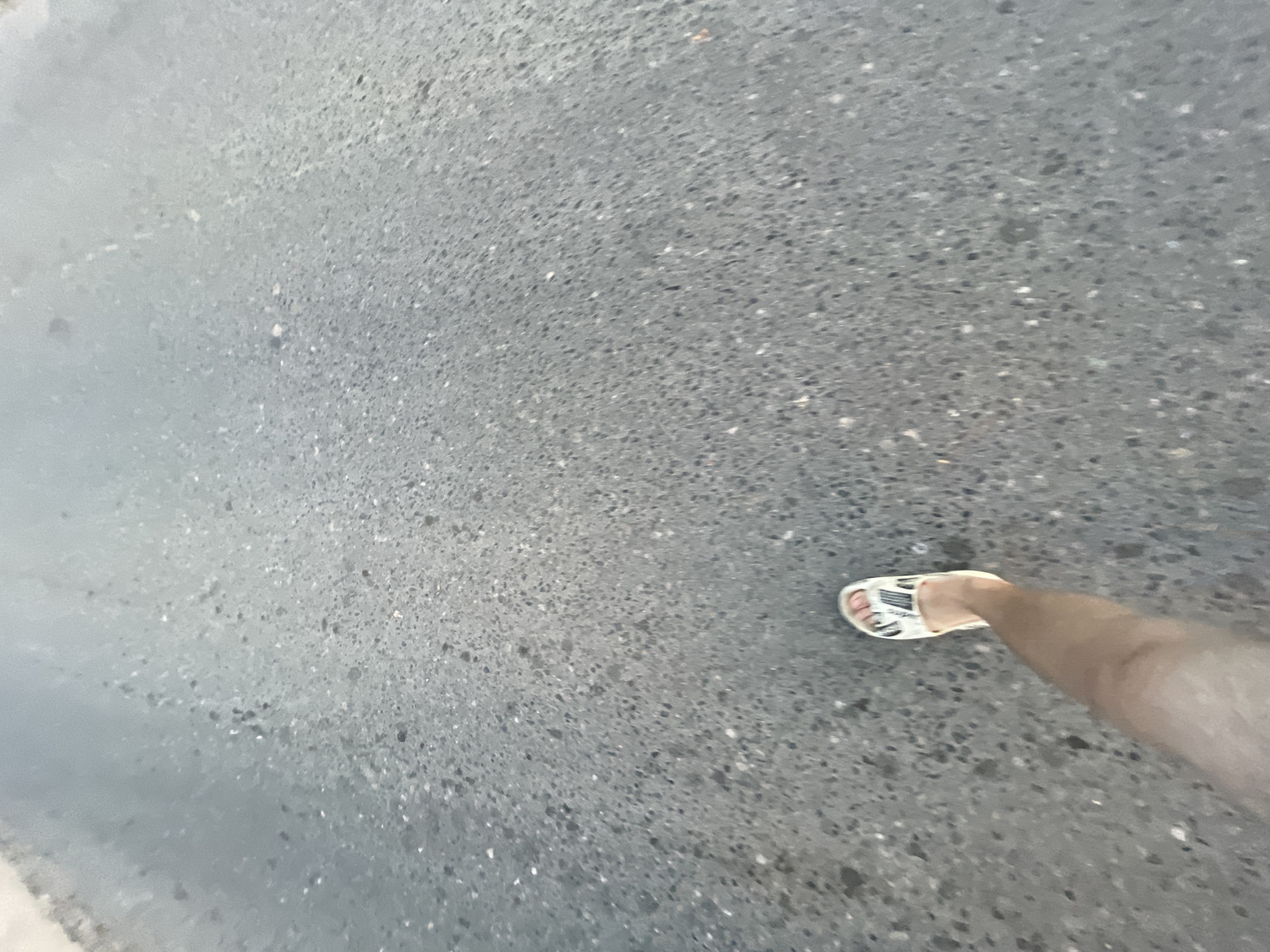 The image shows a first-person perspective of a person's legs and feet walking on an asphalt road. The right leg is visible from the thigh down, wearing a white slide sandal with a black and white patterned strap. The left foot, partially visible, is also wearing a similar sandal. The asphalt surface is dark gray with visible aggregate. The scene appears to be outdoors, with the road stretching into the distance. There are no other people or objects clearly identifiable in the frame, nor any specific landmarks that would indicate the location in Krasnye Baki, Russia. The lighting suggests it is daytime.