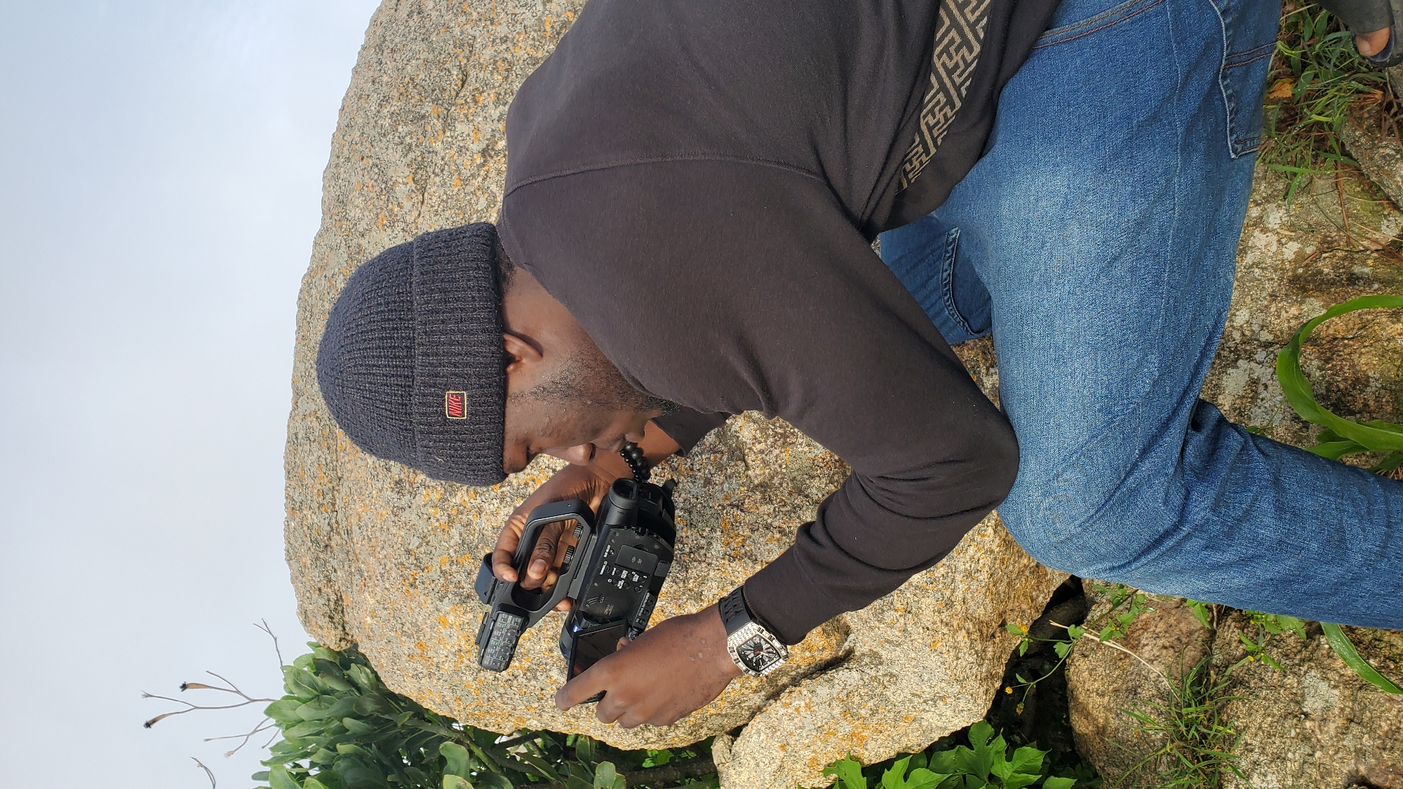 An adult male is crouched on rocky terrain, holding a camcorder and looking through the viewfinder. He is wearing a dark beanie with a "NIKE" logo, a black long-sleeved shirt, and blue jeans. A watch with a black band and a metallic square face is visible on his left wrist. The background consists of a large rock formation, sparse green vegetation, and a pale, overcast sky. The location is likely outdoors, possibly in a natural or semi-natural setting within the city of Jos, Nigeria, characterized by its rocky outcrops and highland vegetation.